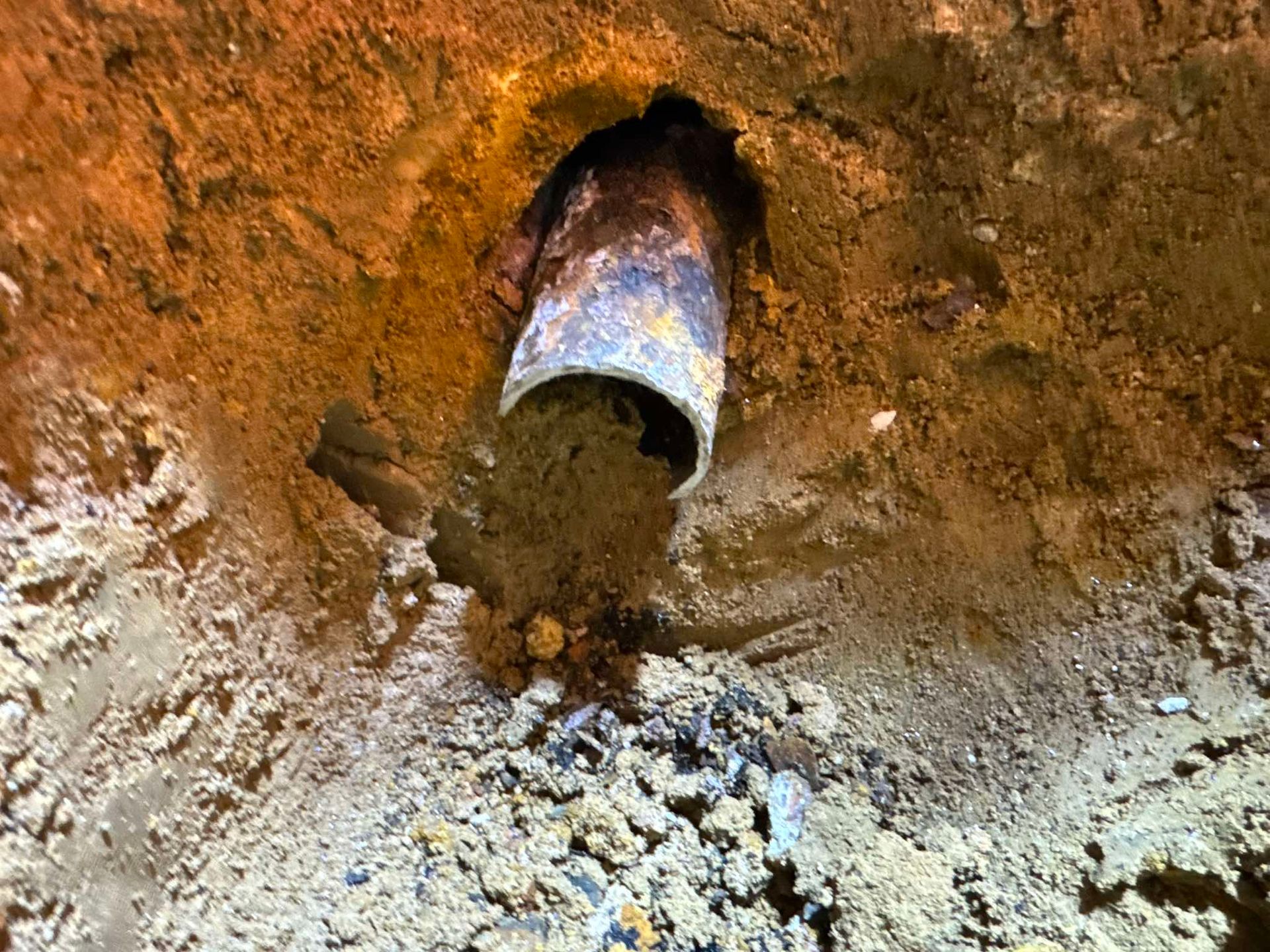 A broken, dirt-filled pipe protrudes from a hole dug into the orange, rocky soil of a trench.
