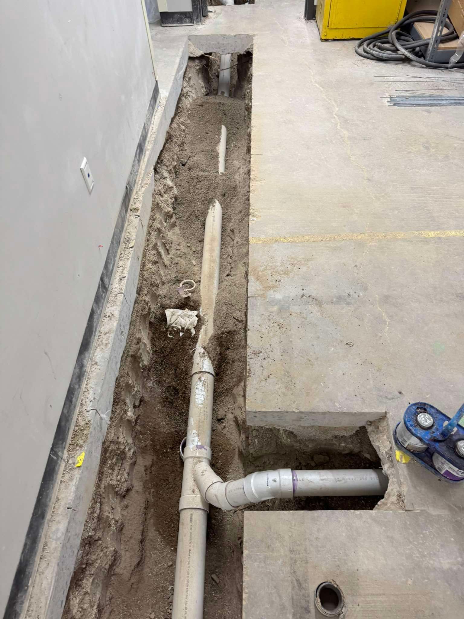 A view of a trench cut into a concrete floor, revealing exposed plumbing pipes and soil during a construction project.