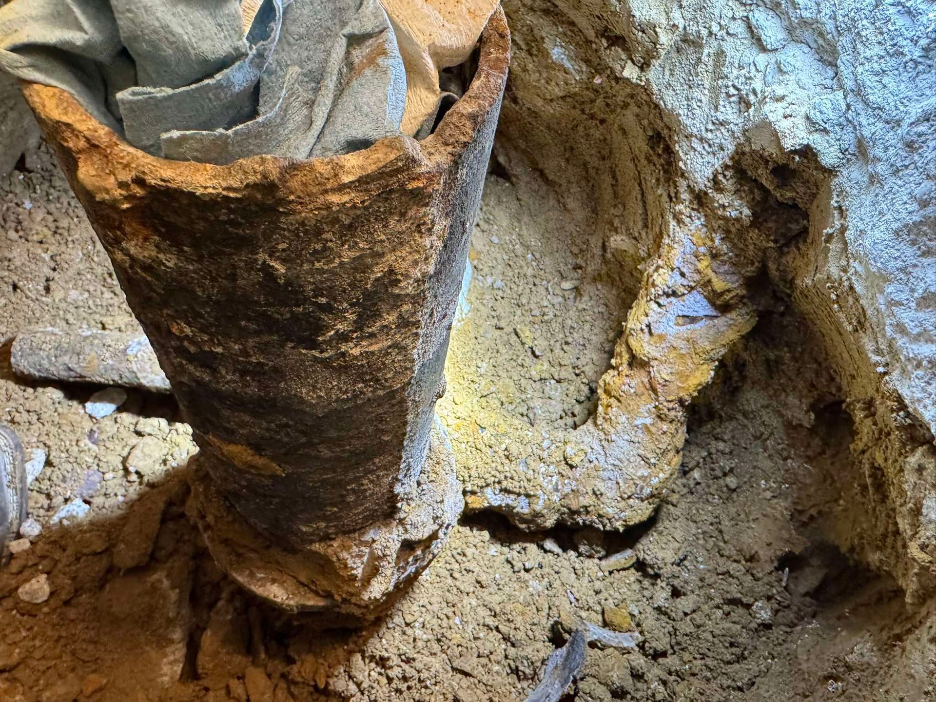 A corroded cast iron drain pipe opening with a gray rag inside, set within an excavated dirt and concrete floor.