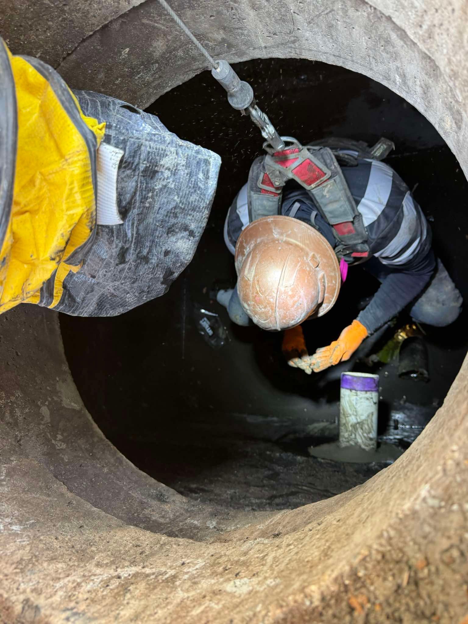 A person in a hard hat and safety harness works inside a dark, concrete circular shaft while attached to a safety line.