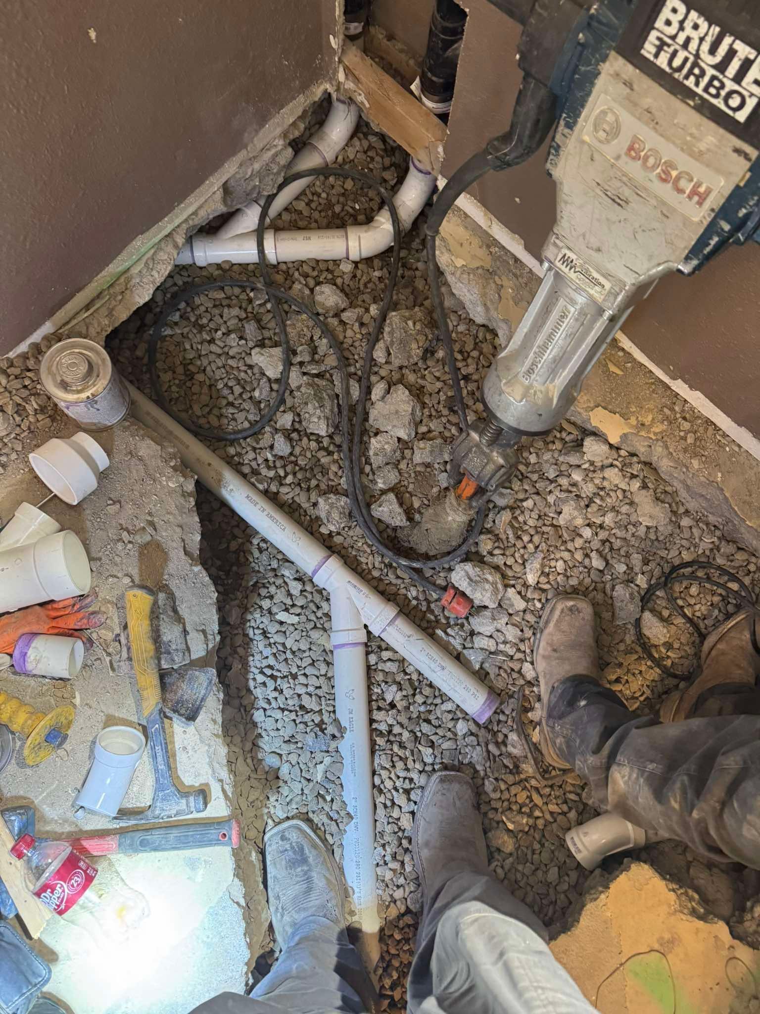 A construction worker uses a Bosch jackhammer to break up a concrete floor, revealing plumbing pipes underneath.