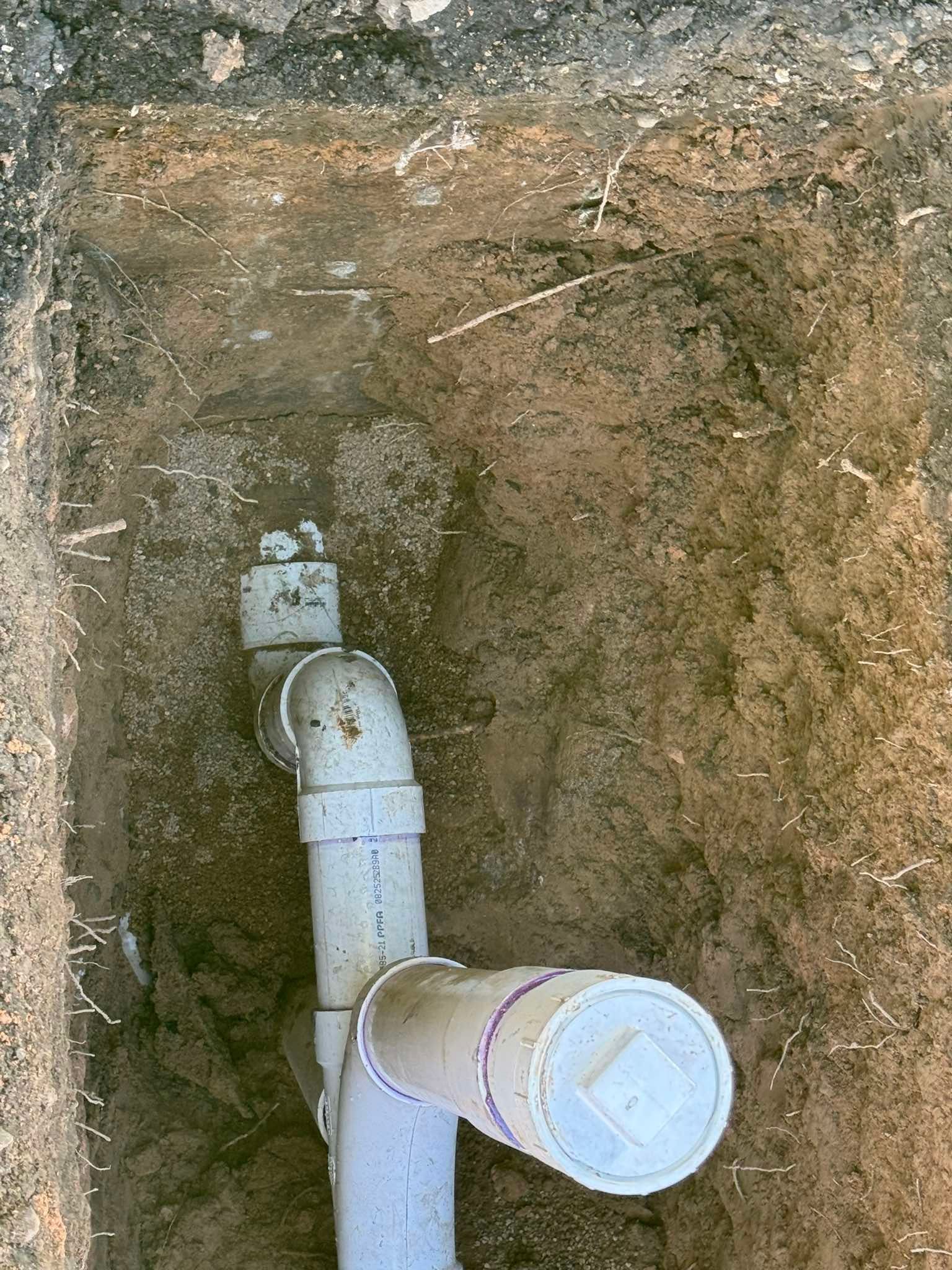 A light gray PVC drain pipe with a capped vertical cleanout fitting partially buried in a dirt trench.