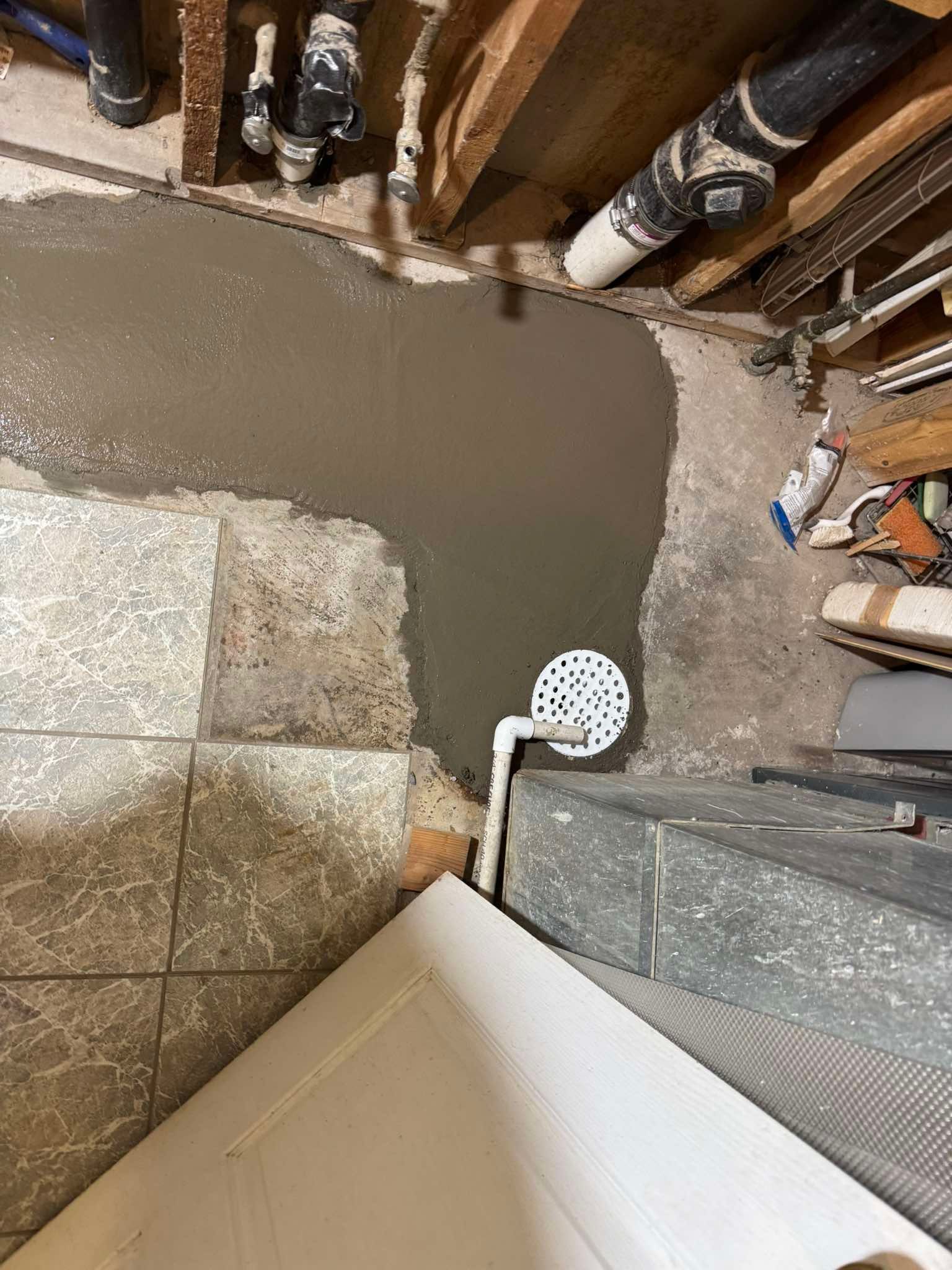 A patch of wet concrete floor with a white circular floor drain, situated near tiled flooring and exposed wall studs.