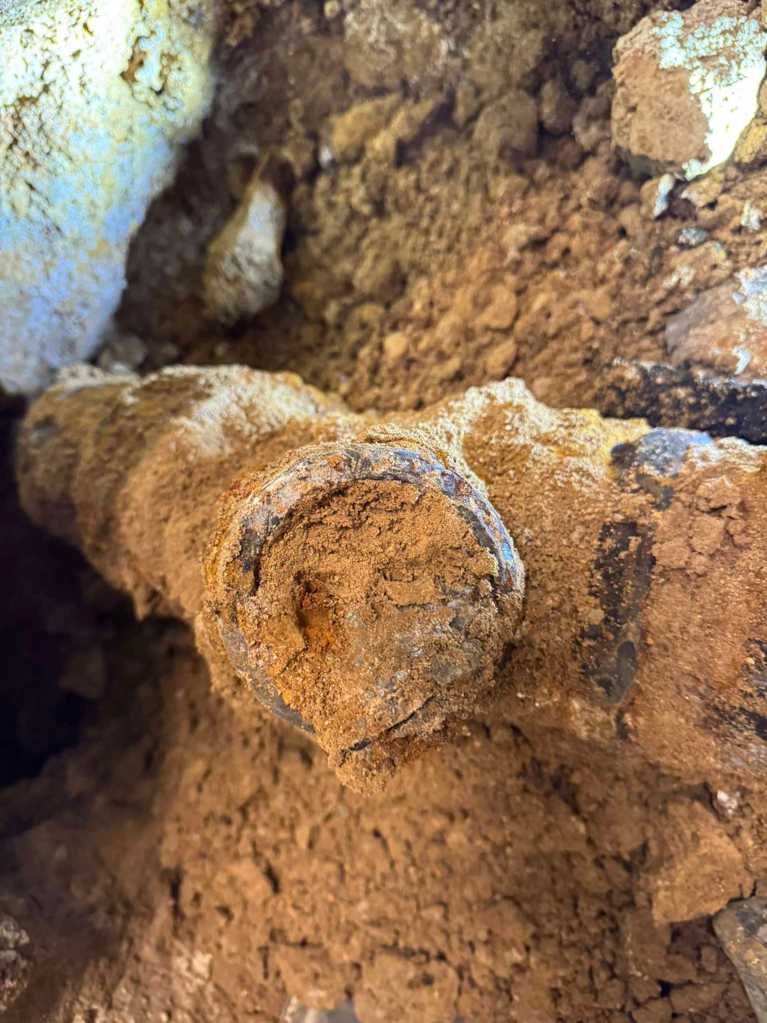 A section of rusted, soil-covered metal pipe is exposed in a trench, showing a blockage inside.