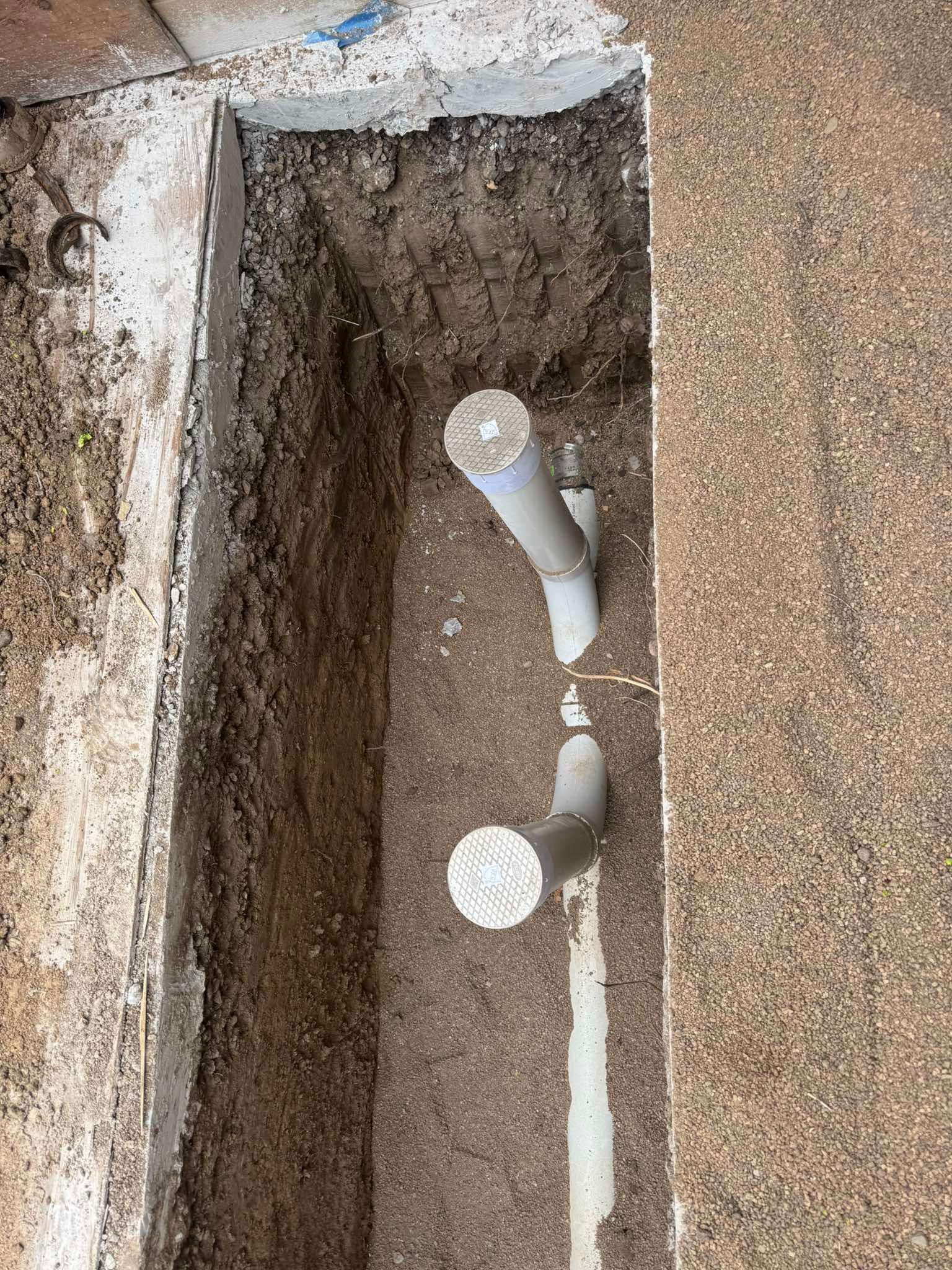 Two gray PVC plumbing pipes protrude vertically from a dirt trench cut into a concrete floor.