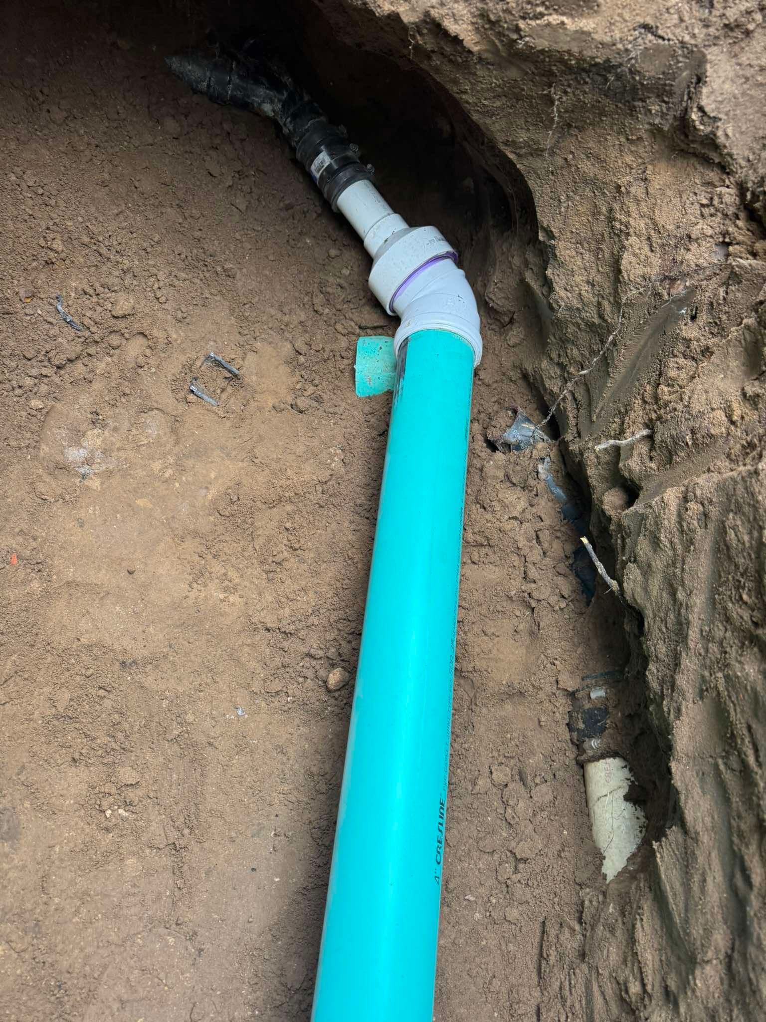 A blue irrigation pipe connects to a white PVC elbow and adapter in a trench dug into brown soil.