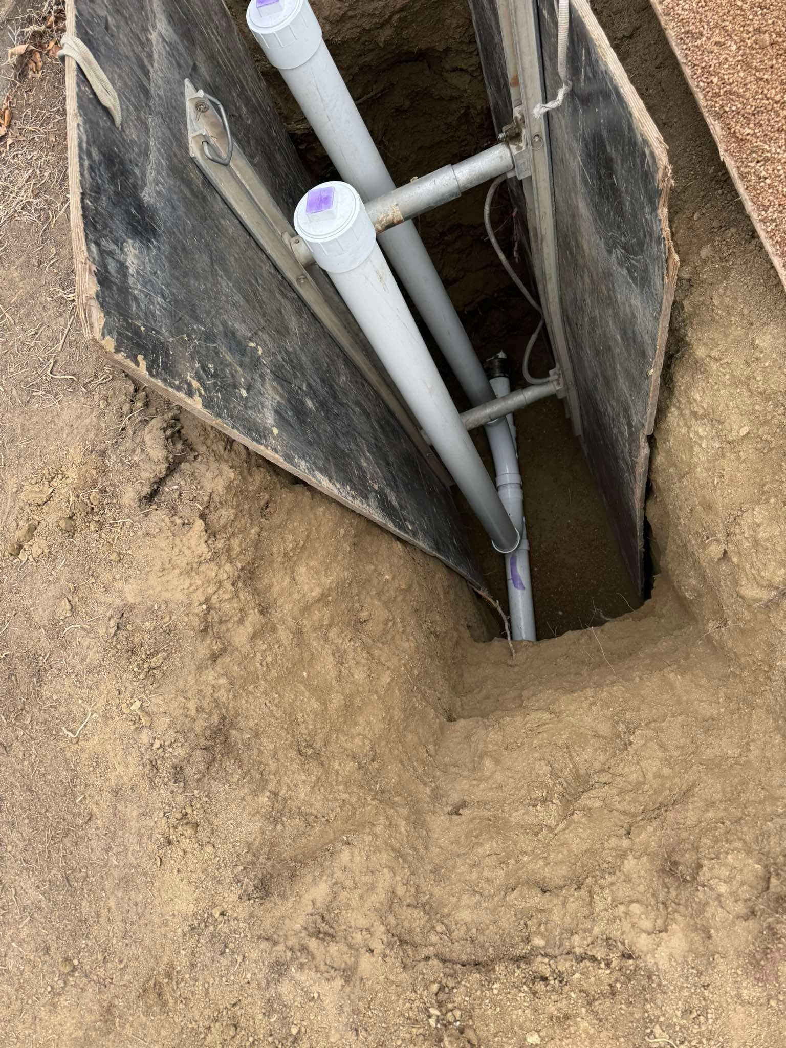 A high-angle view of two grey PVC pipes positioned within a narrow, earth-walled trench supported by metal bracing.