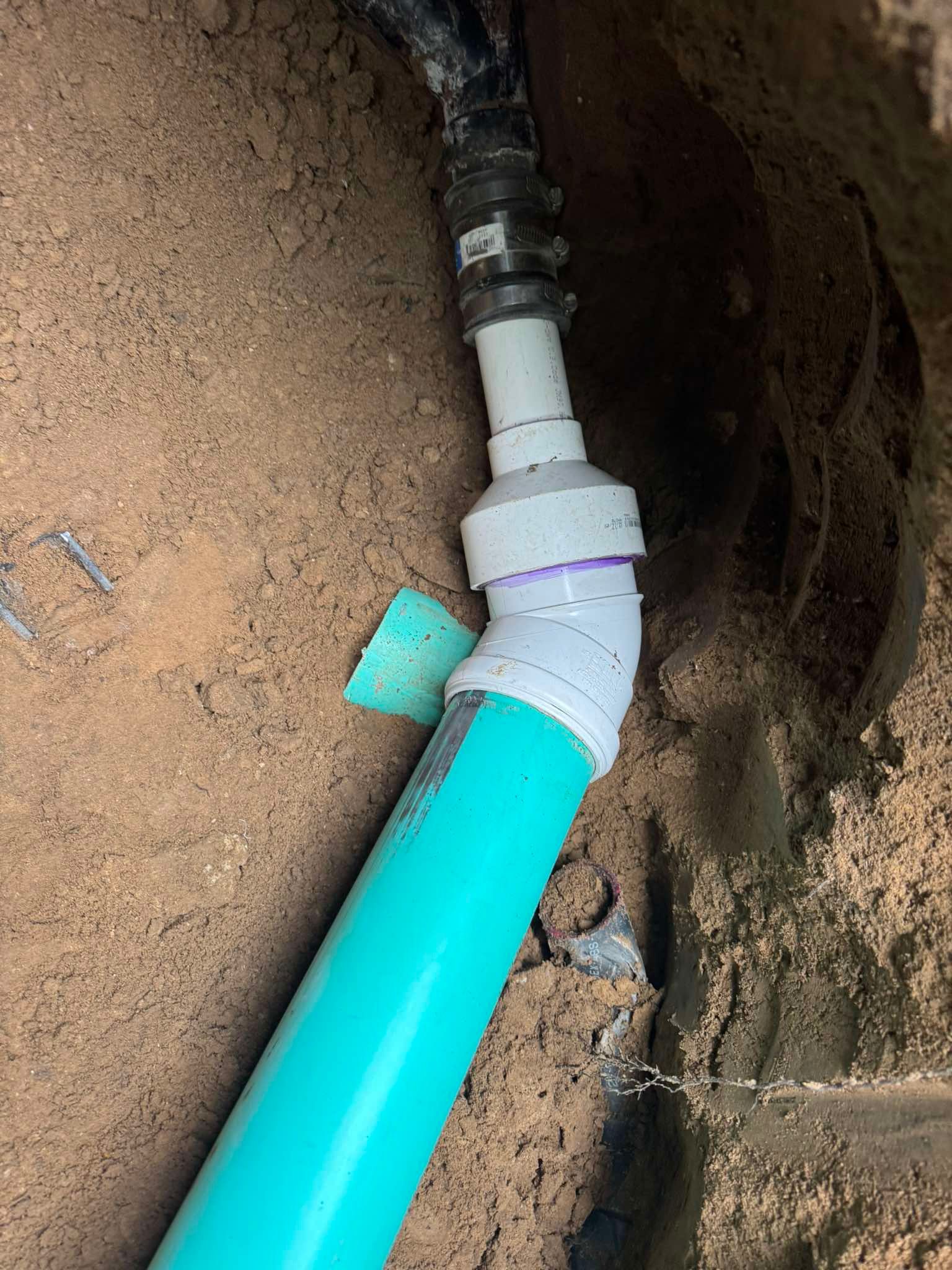 A PVC pipe connection in a trench, showing a green pipe transitioning to white fittings and a rubber coupling.