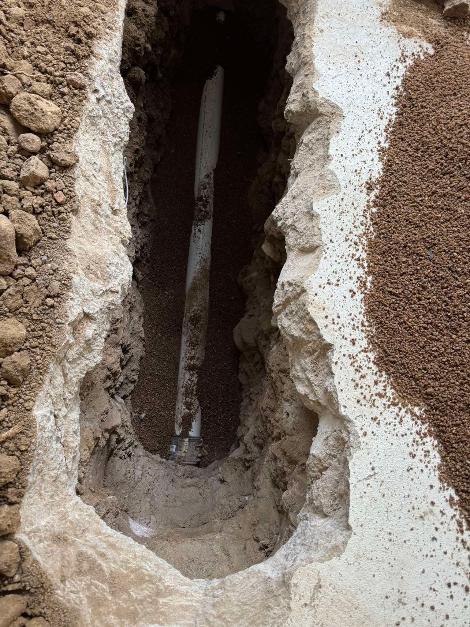 A white pipe is visible inside a narrow, vertical trench dug into dirt and concrete flooring.