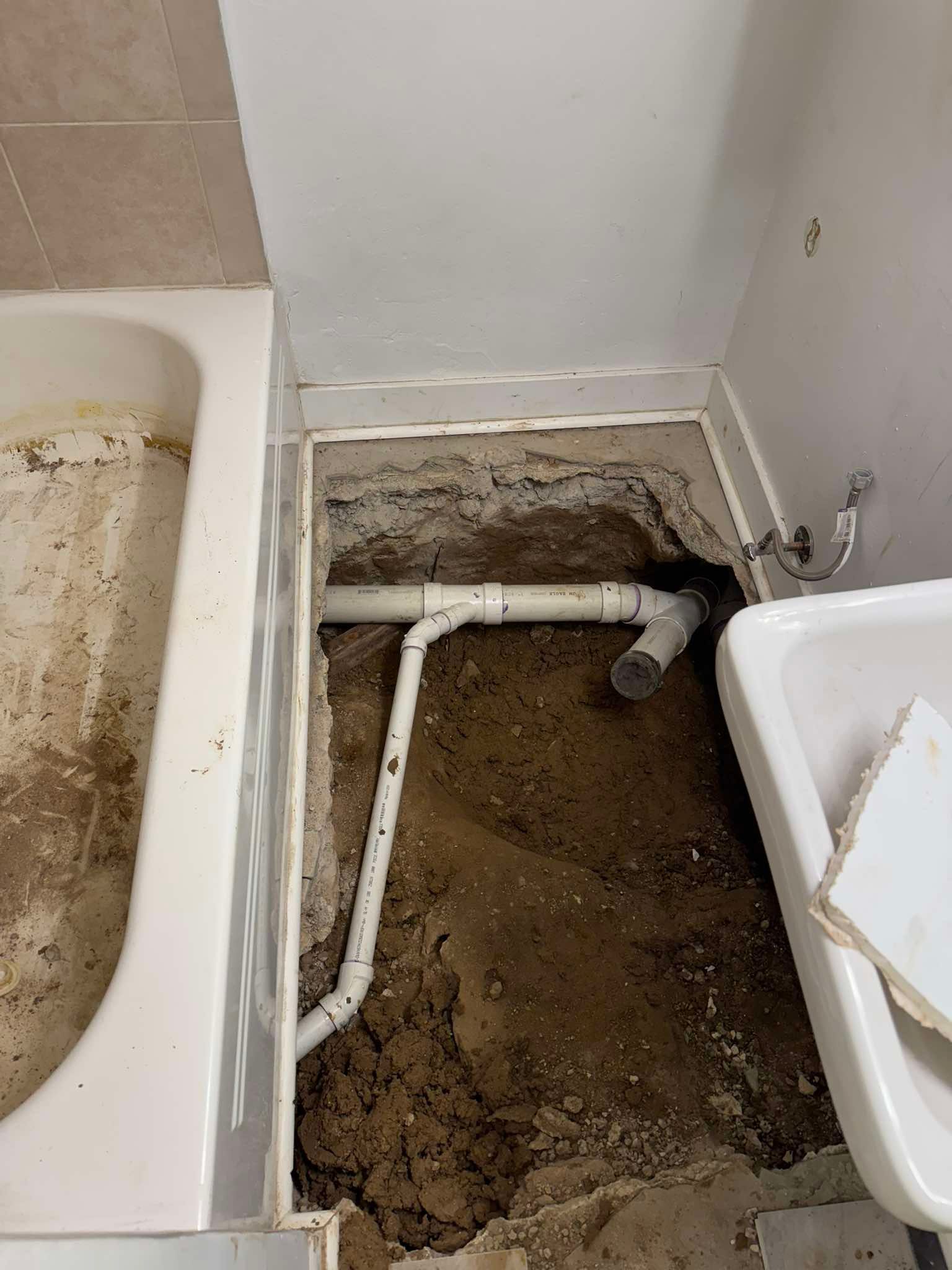 A bathroom floor with an open trench revealing exposed white PVC plumbing pipes connected to a tub and sink drain.