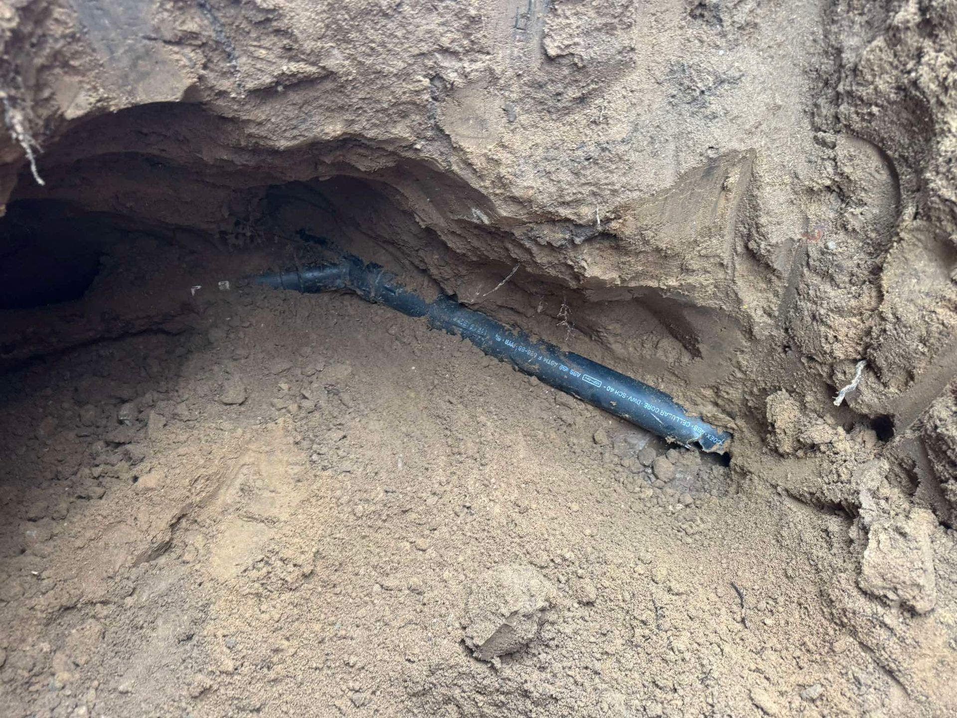 A black plastic utility pipe lies exposed at the bottom of a freshly dug, sandy dirt trench.