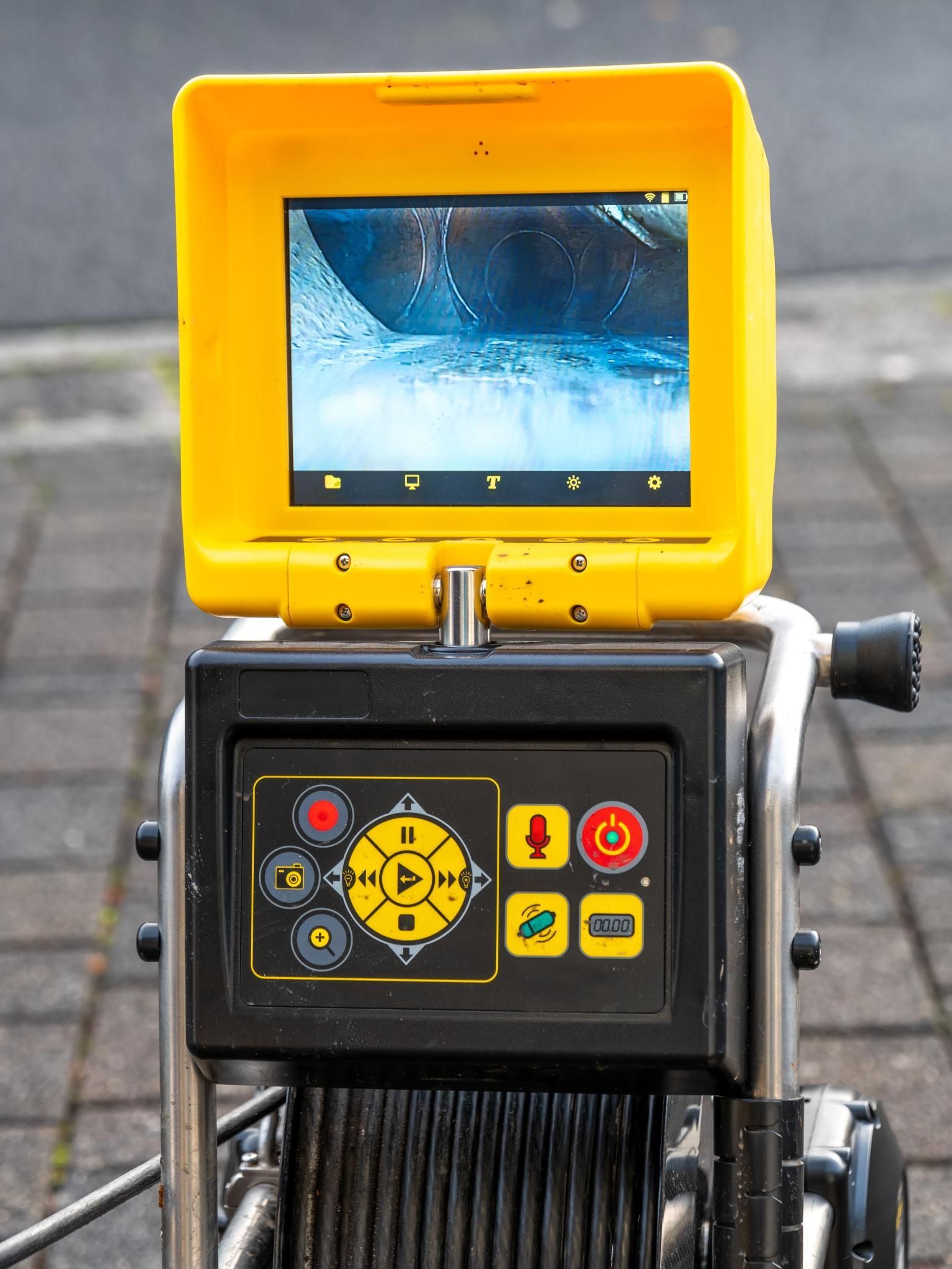A yellow, portable pipe inspection camera system with a screen displaying an interior view, sitting on a paved surface.