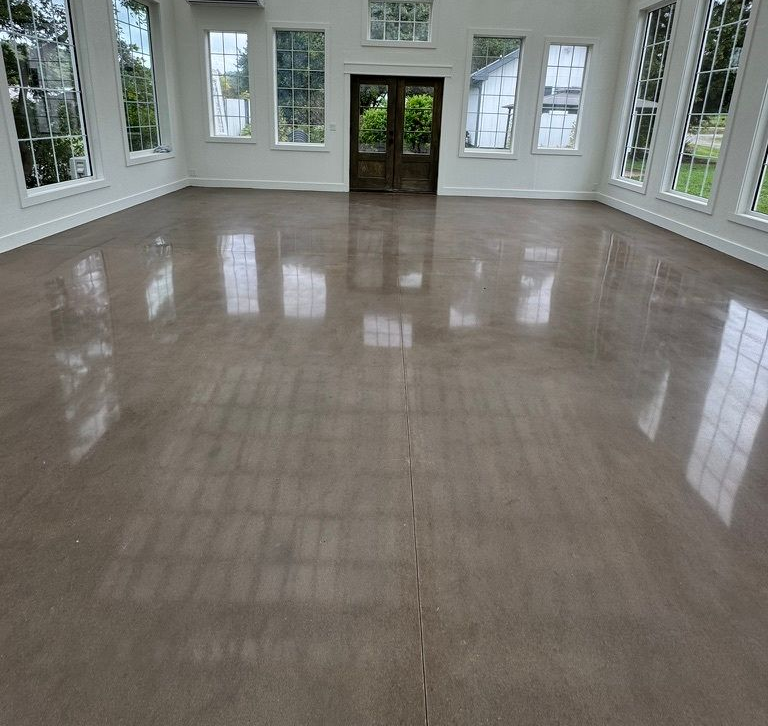 A bright, empty room with white walls, large multi-pane windows, and a highly polished, reflective concrete floor.
