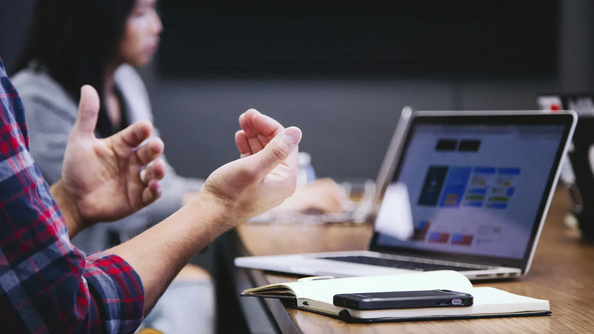 A person gesturing during a meeting, laptop and notebook on the table, another person in background