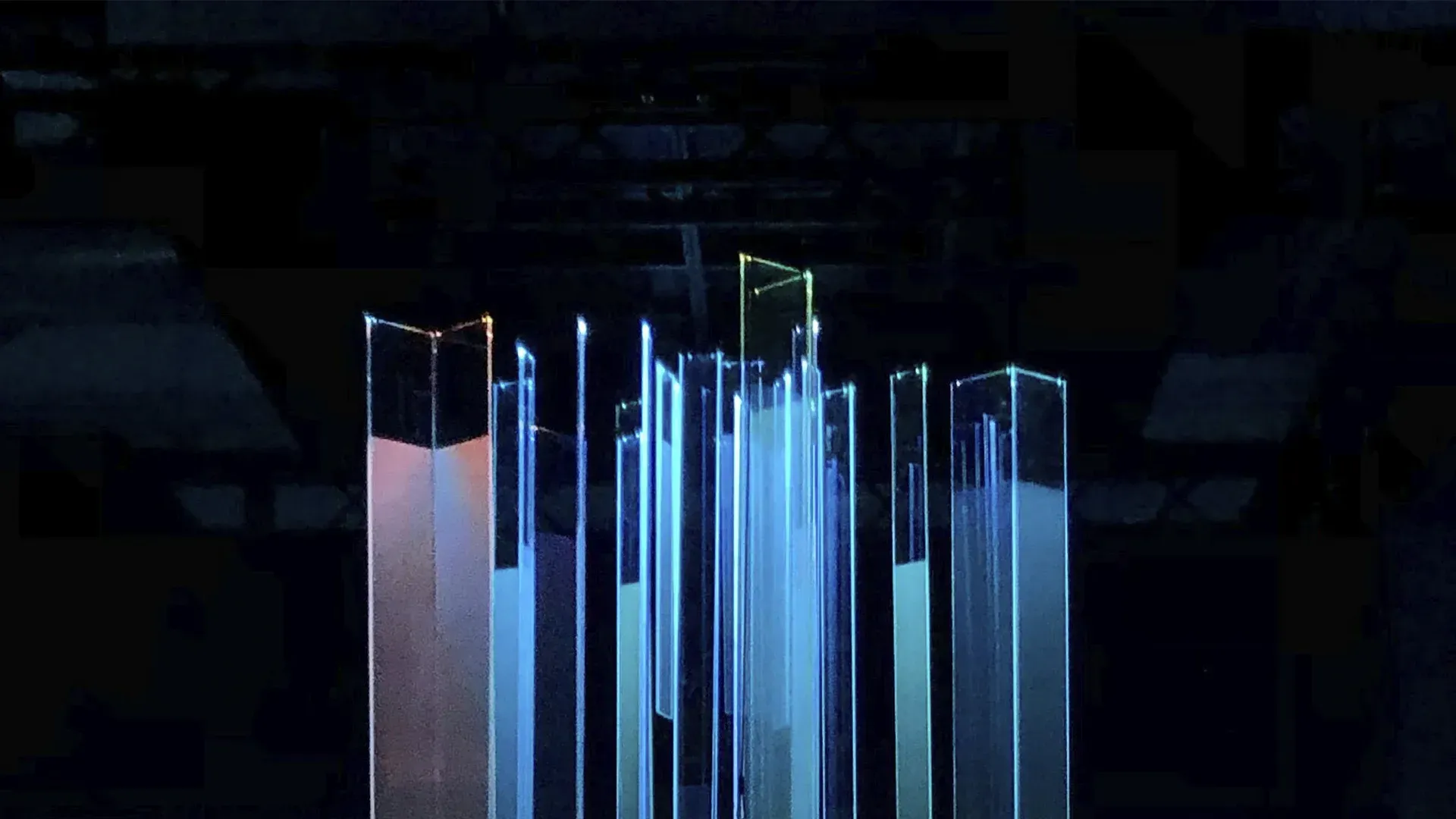 Abstract cityscape of blue-lit rectangular structures, some with orange and red hues. Dark background.