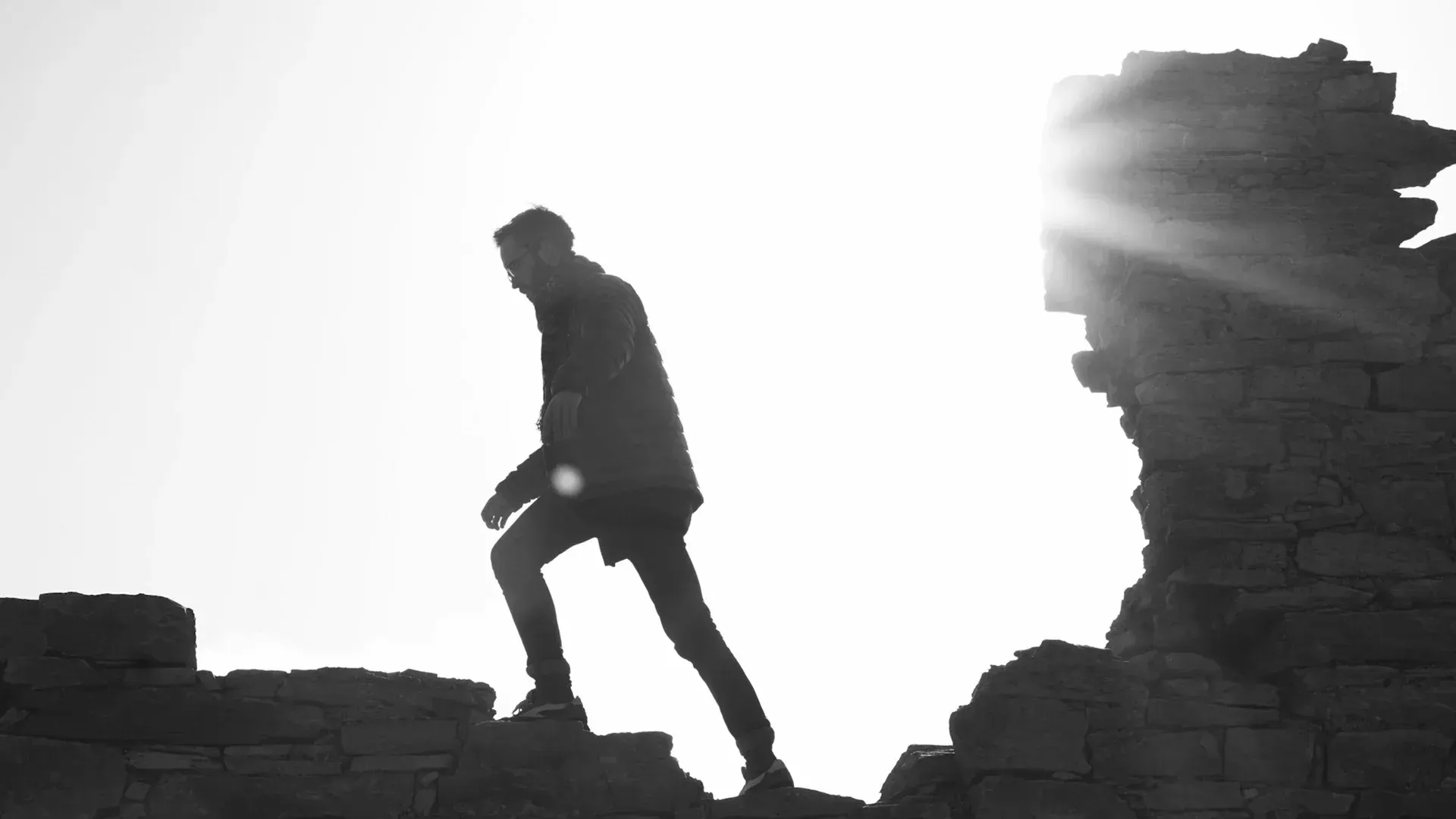 Silhouette of a person climbing over a stone wall with the sun shining brightly behind them.