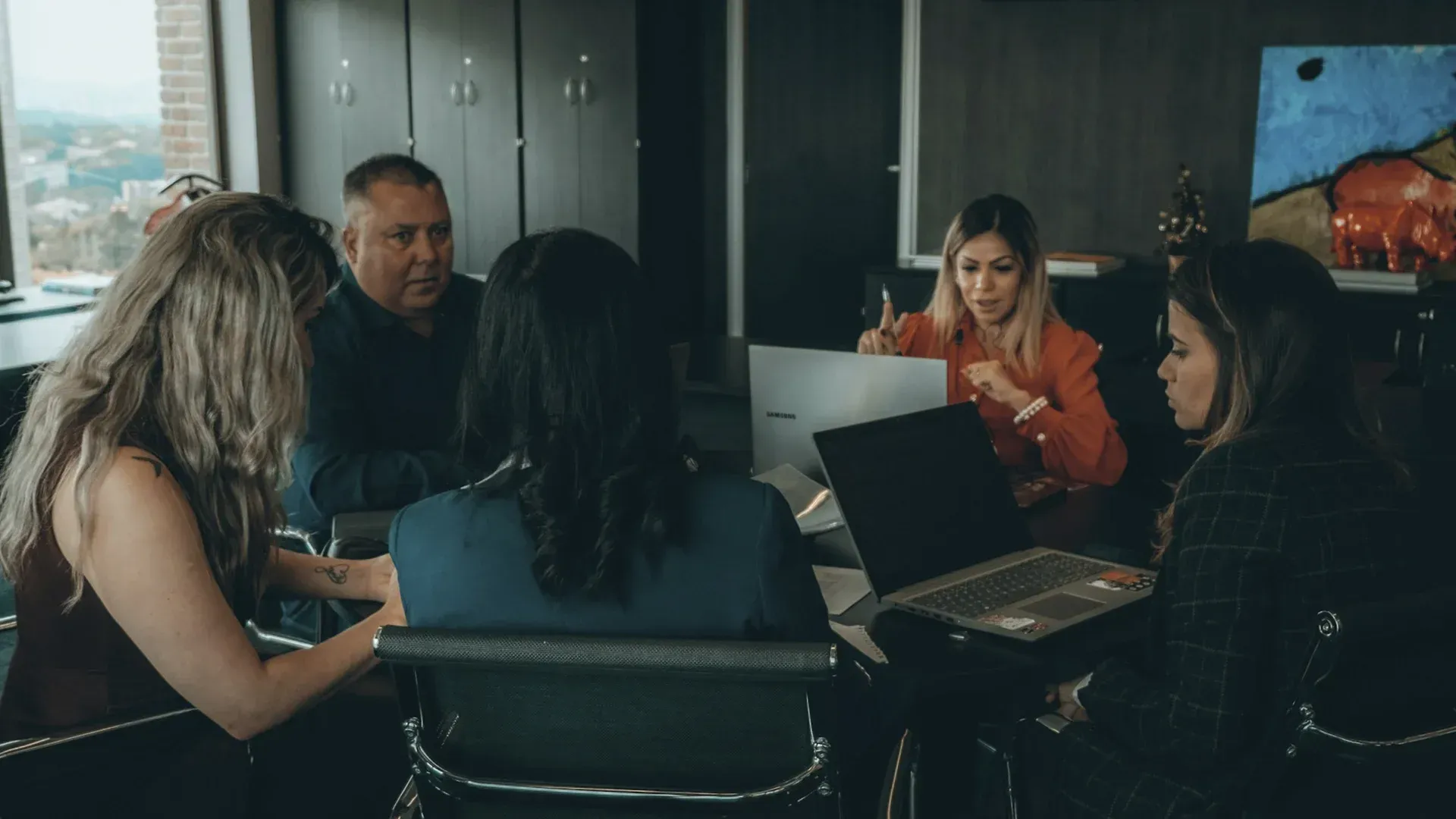 People gathered around a table with laptops, one woman in red points while others observe in a modern office.