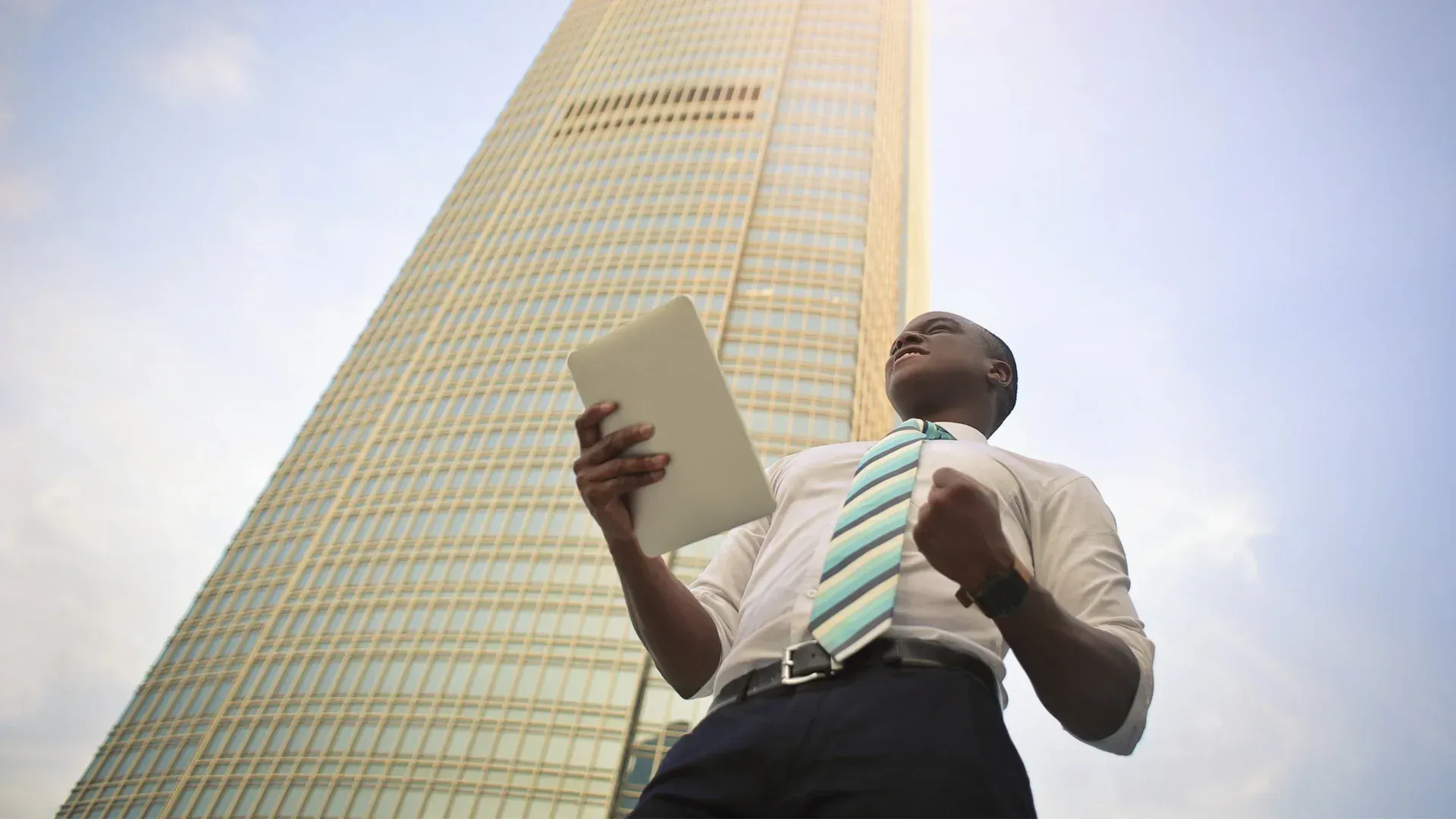 Man with tablet, fist raised in front of a tall skyscraper, celebrating a success.