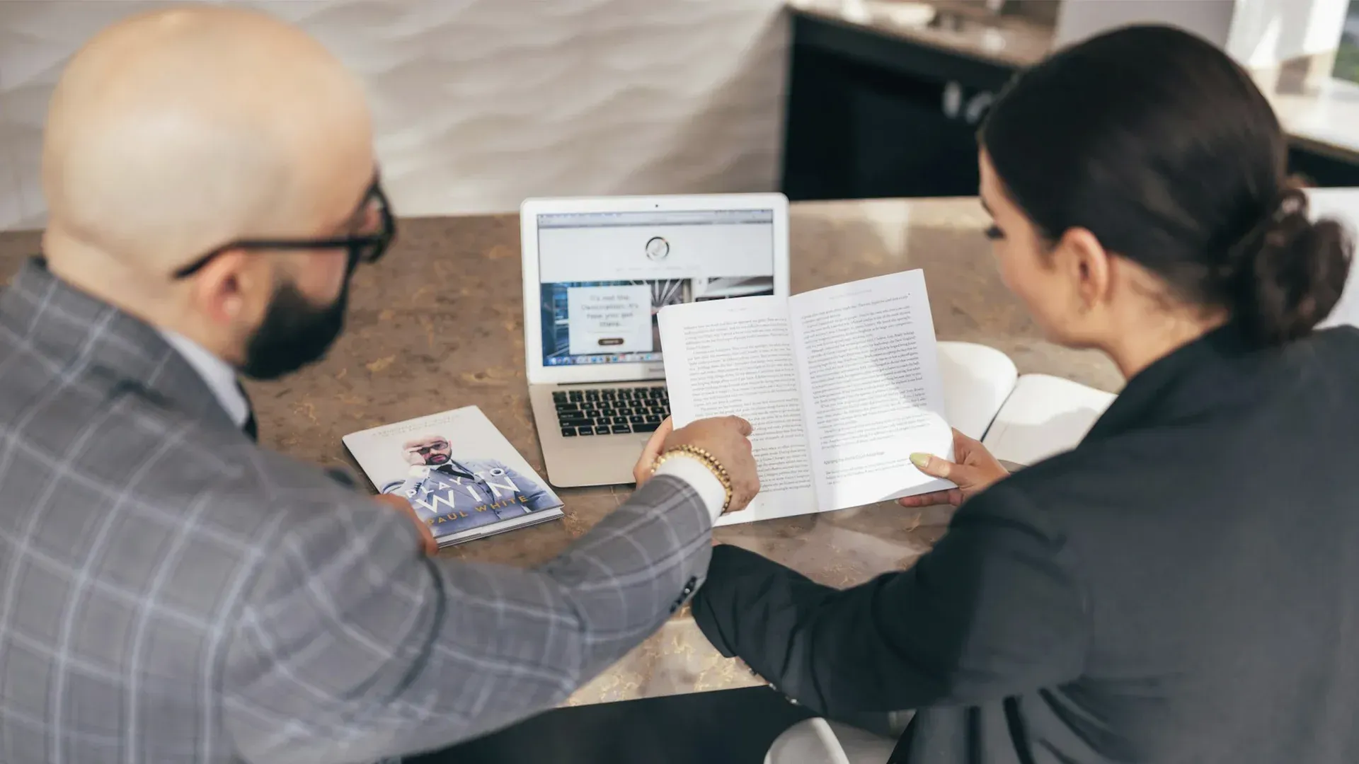 Two people reviewing documents, a laptop open on the counter, a bald man, and a woman wearing a blaz