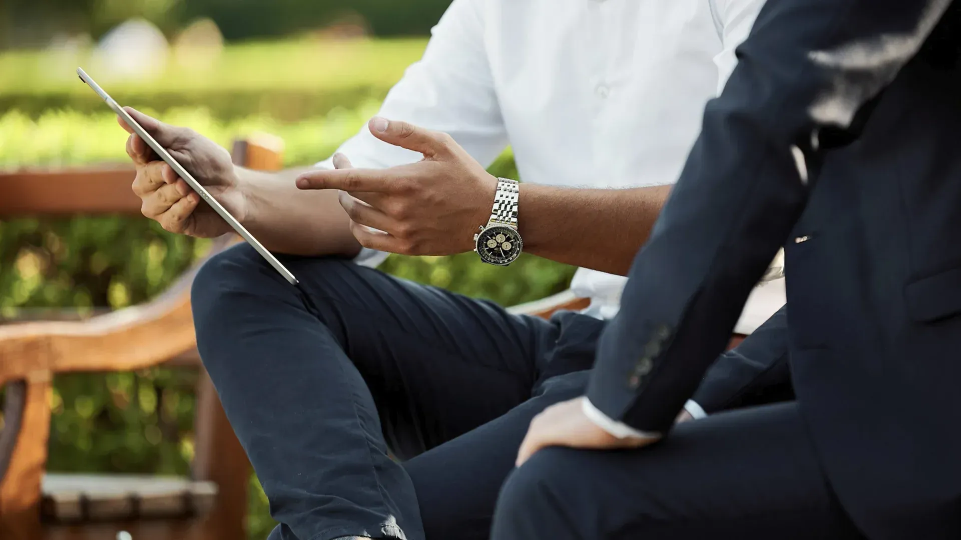 Two men in business attire, one holding a tablet and gesturing, sitting on a bench outdoors.