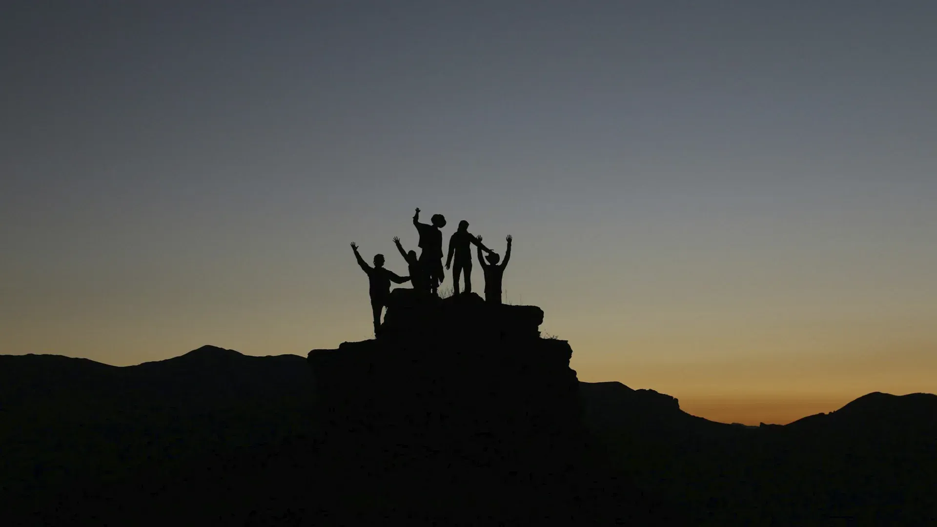 Silhouetted figures with arms raised, atop a mountain, against a dusk sky.