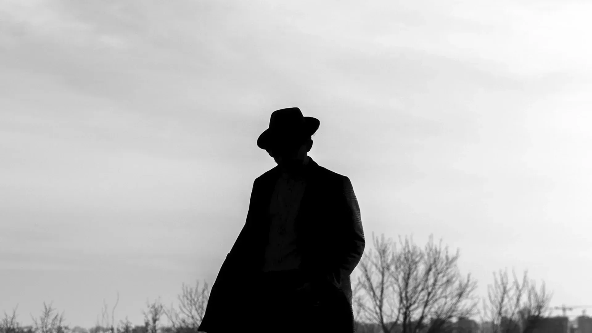 Silhouette of a person in a fedora and coat, standing outdoors.