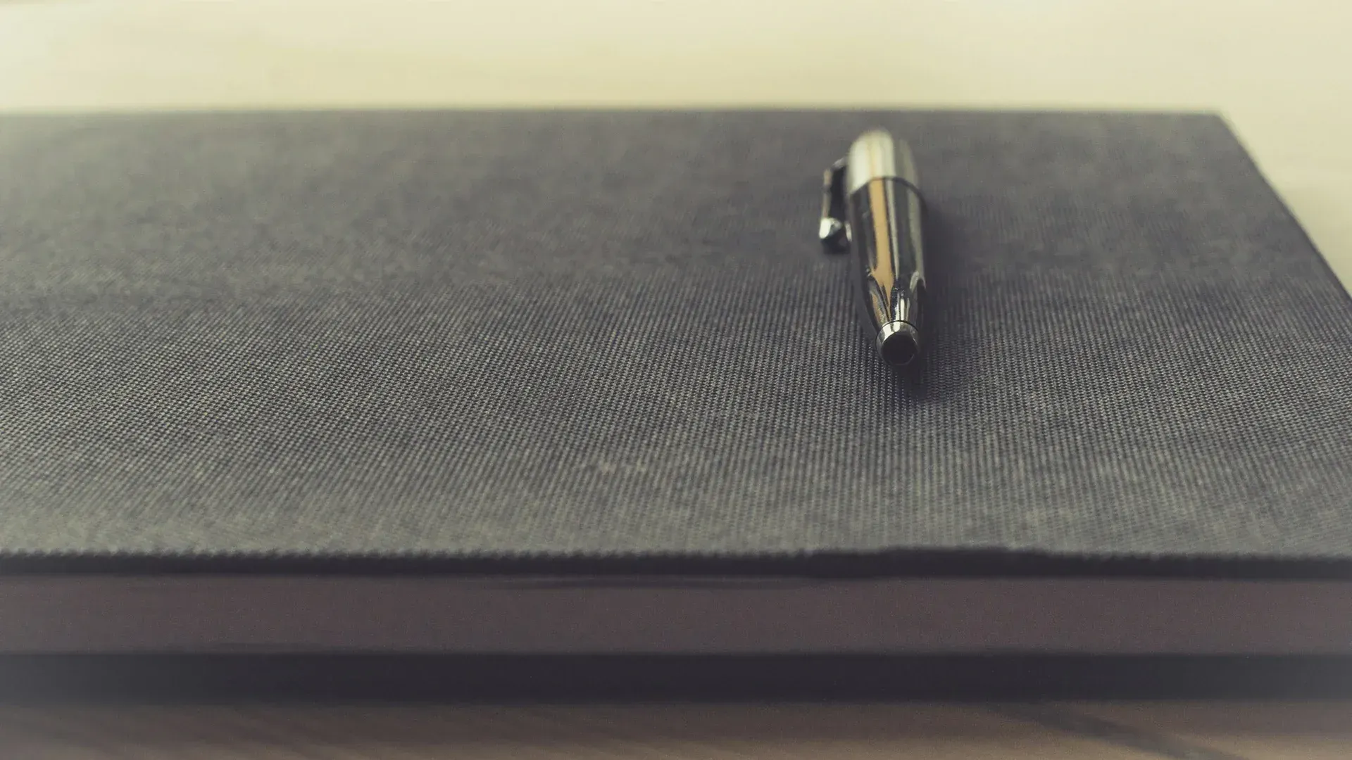 Black notebook with a silver pen on top, resting on a light wooden surface.