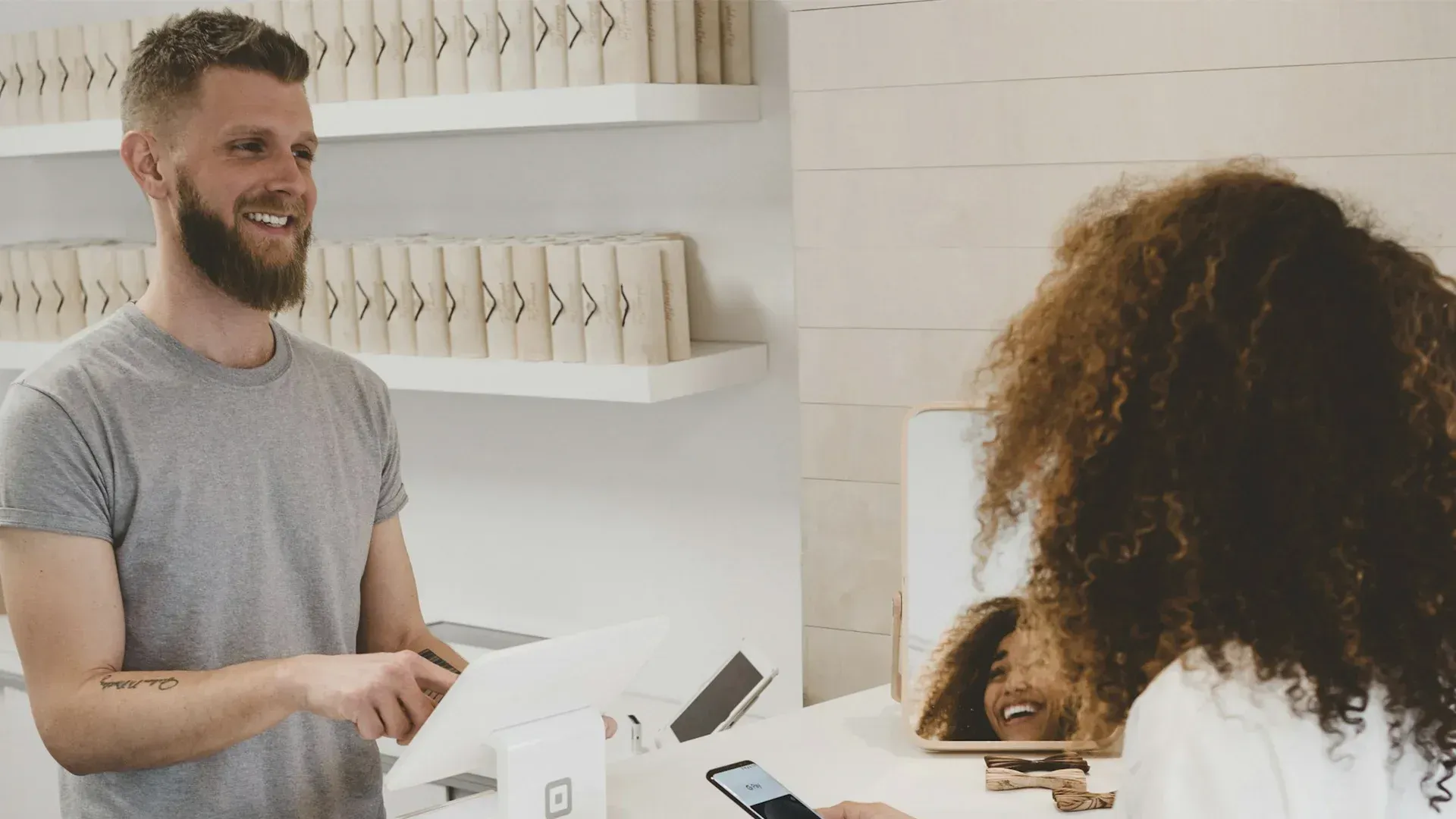 Man behind counter smiles, assisting a customer. Customer's smile is reflected in a mirror. White interior.