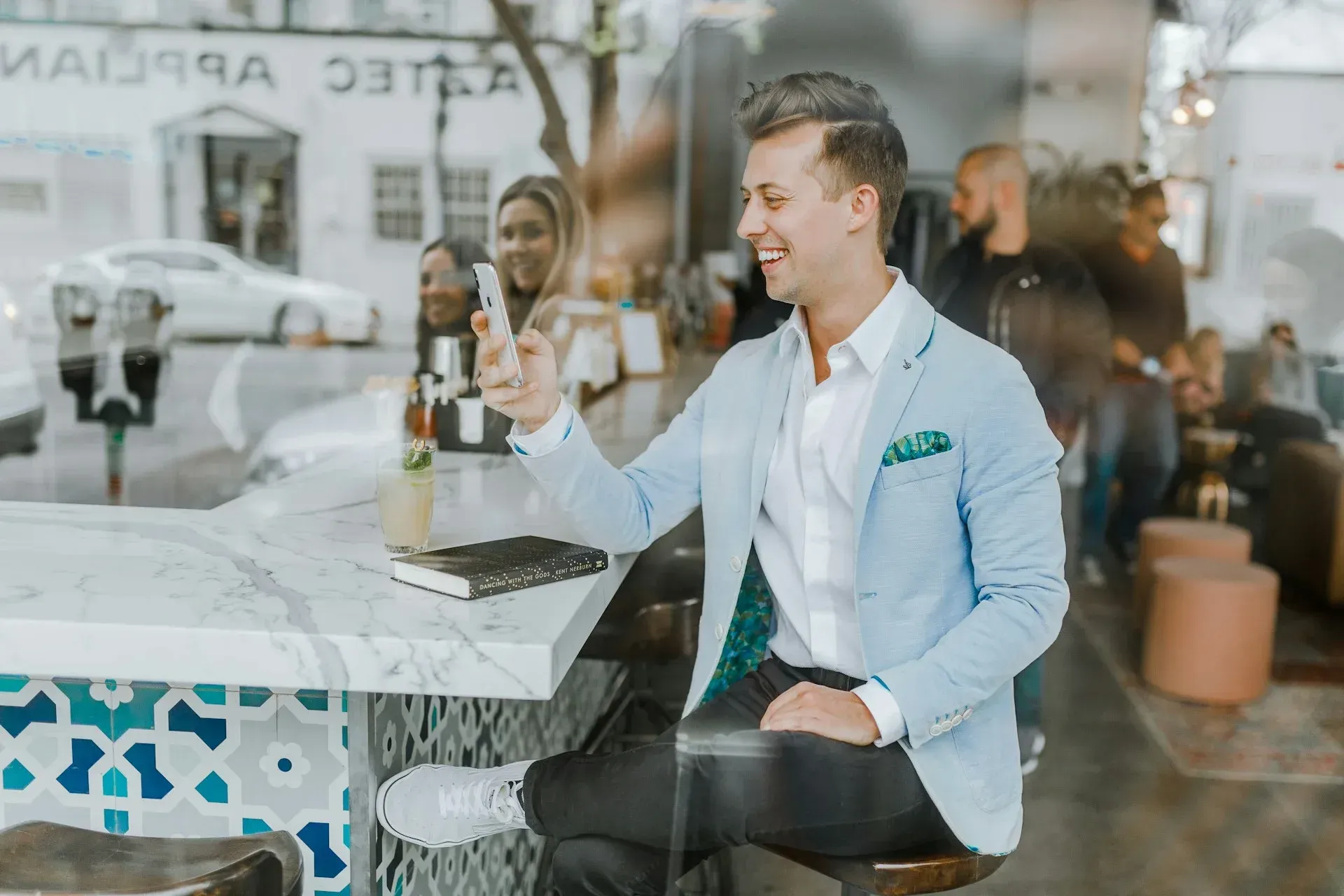 Man in blue blazer smiles at phone, seated at a bar with a patterned front.