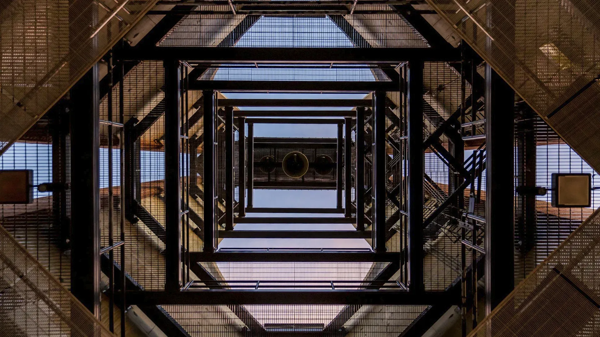 Looking up through a geometric steel framework, towards a decorative ceiling, and the sky.
