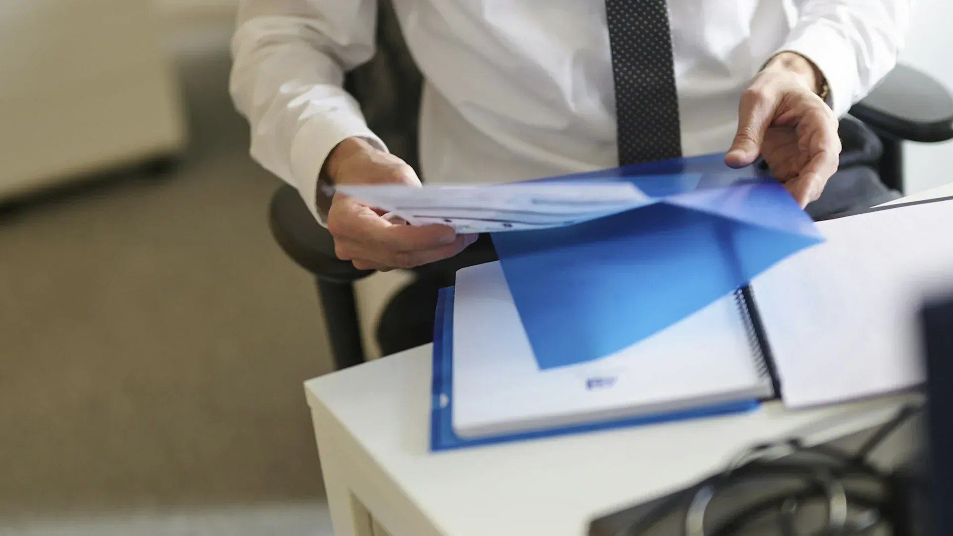 Person in a white shirt and tie reviews documents in a blue folder at a desk.