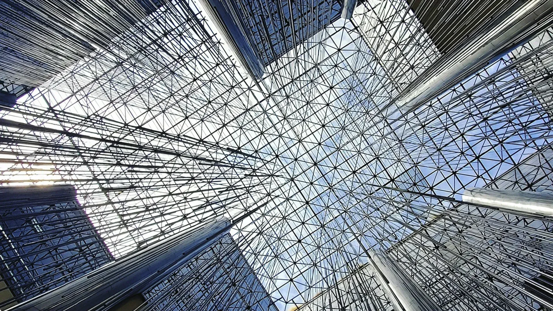 Looking up at a modern glass and steel structure, intersecting beams and a clear sky.