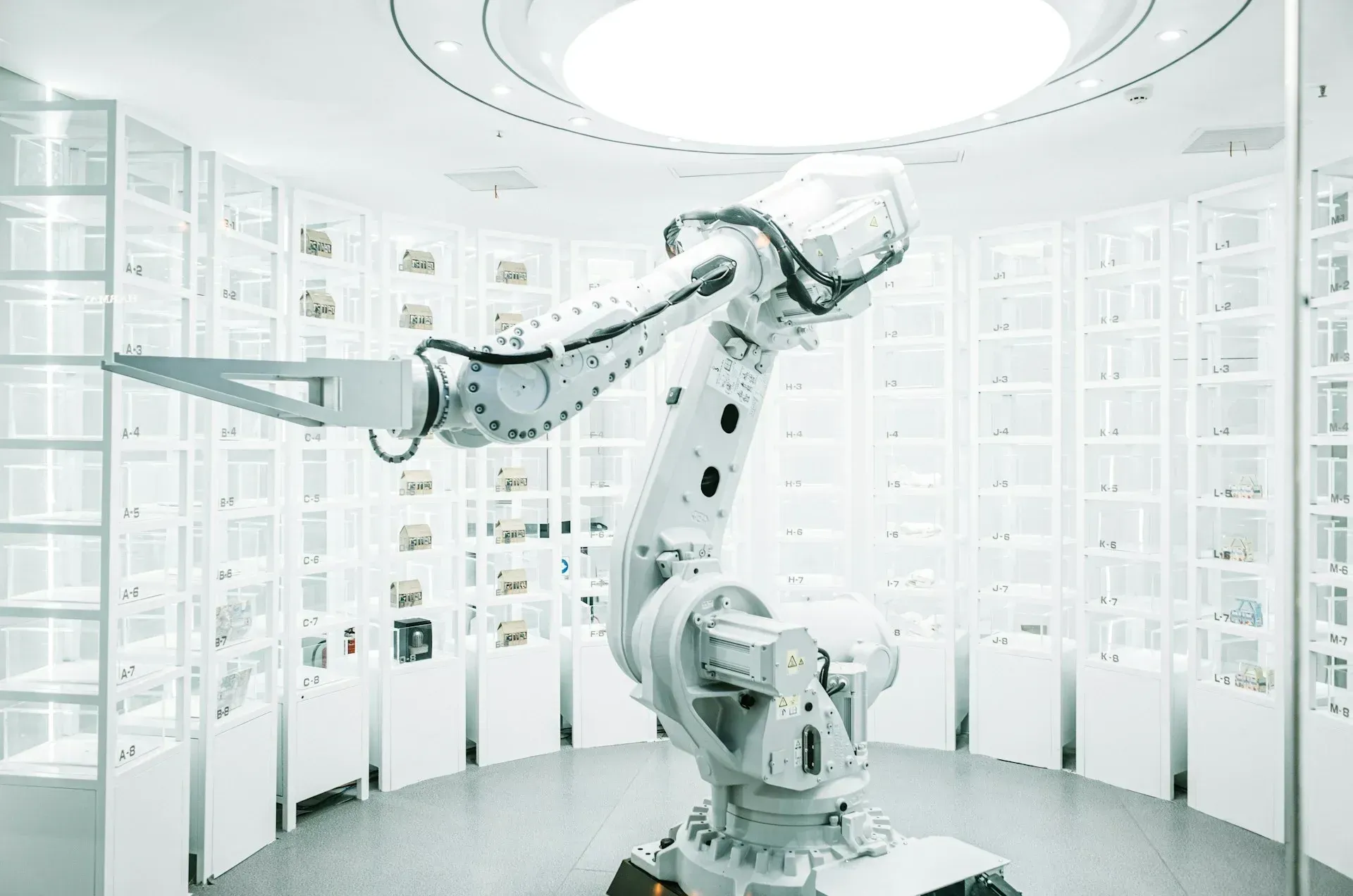 White robotic arm in a circular white room with shelves filled with objects, under a round ceiling light.