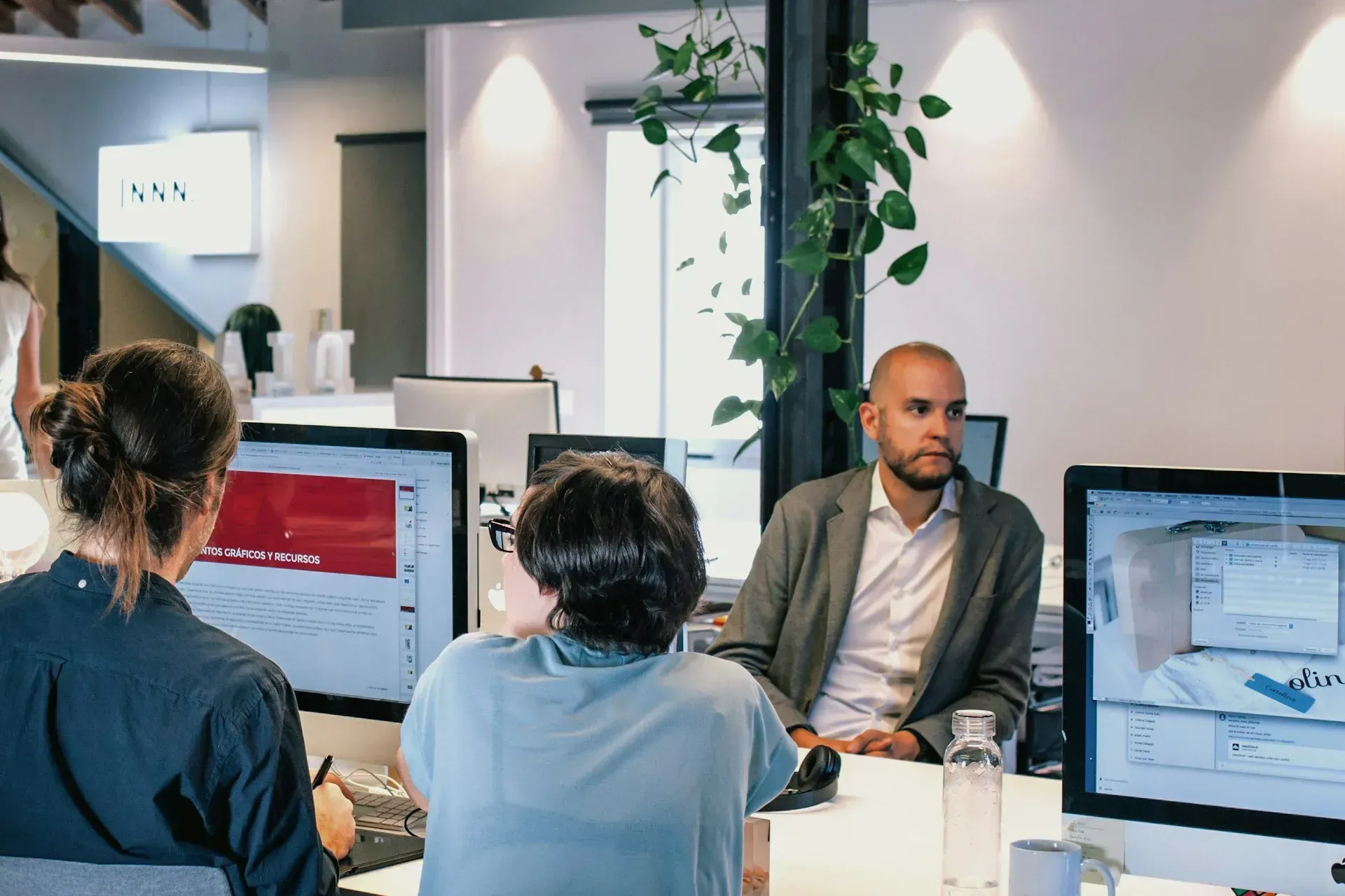 Three people at computers in a modern office; a man in a blazer is listening.