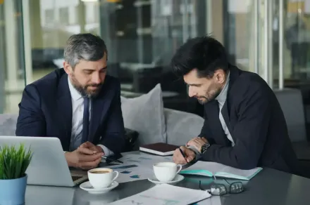 Two men in suits reviewing documents at a table with a laptop, coffee, and notepad in an office setting.