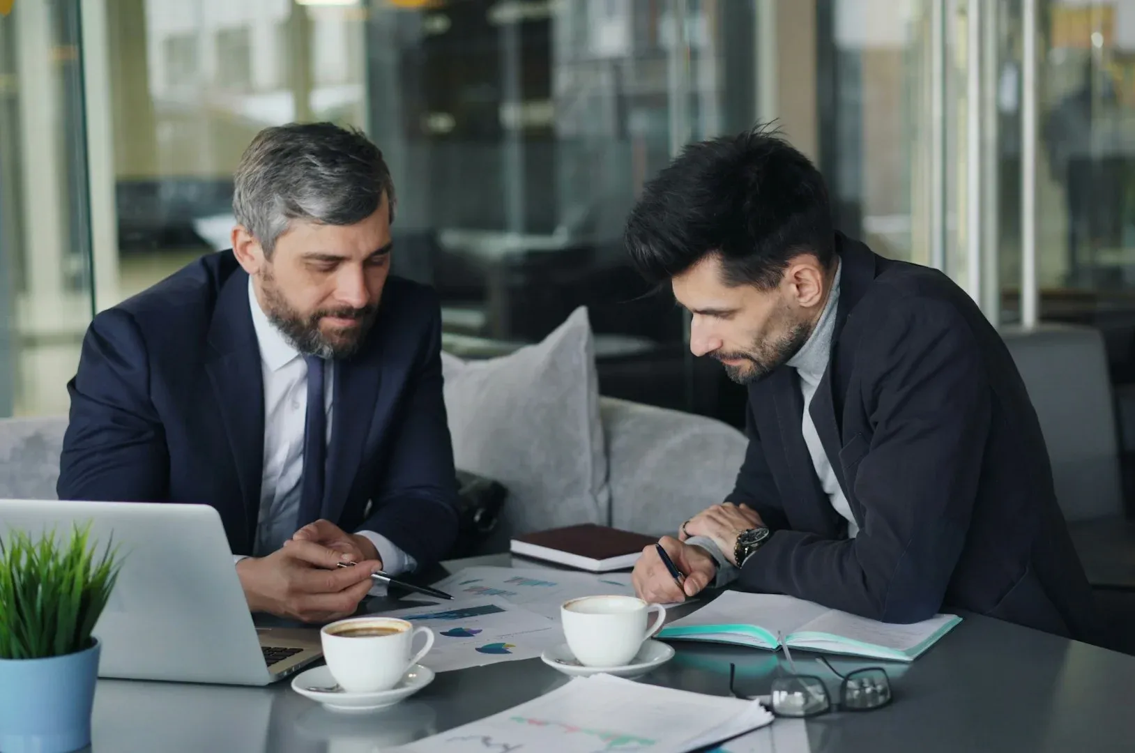 Expert Group Two men in suits reviewing documents at a table with a laptop, coffee, and notepad in an office setting.