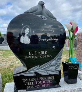 Pierre tombale noire en forme de cœur avec une photo, des colombes et une inscription en français ; à côté, un pot de fleurs.