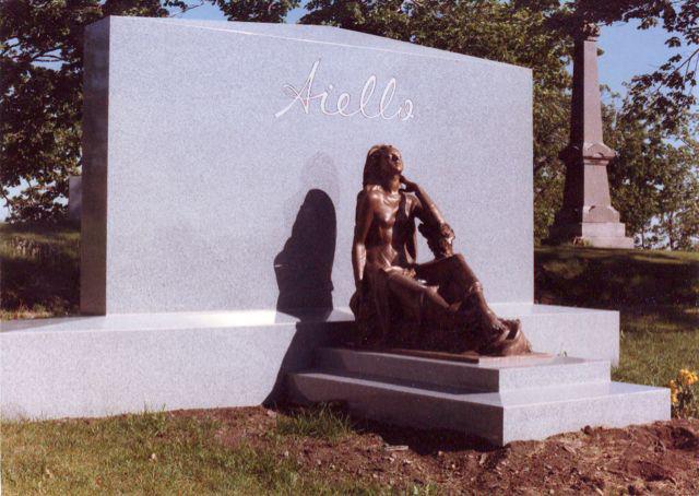 Grand monument gris orné d'une sculpture en bronze représentant deux personnages dans un cimetière. L'inscription « Stolls » figure au sommet.