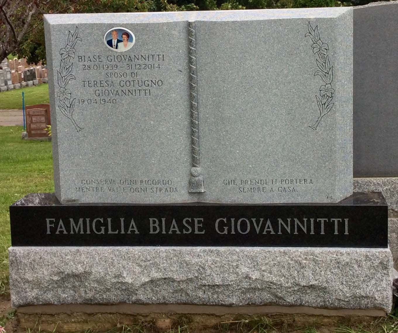 Pierre tombale grise avec noms et dates, motif de livre ouvert et photo. « Famiglia Biase Giovannitti » est gravé sur le socle.