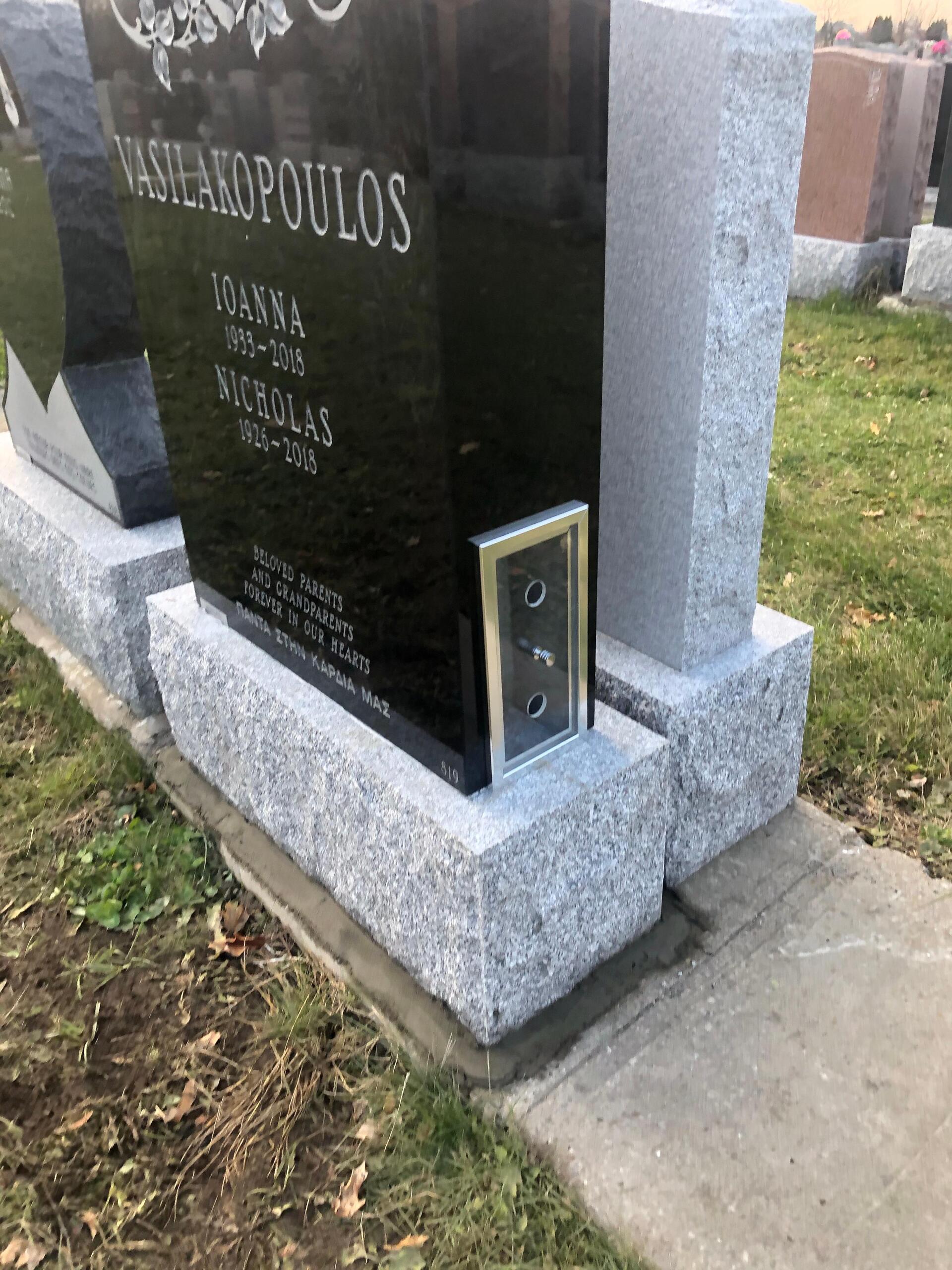 Une pierre tombale noire avec une boîte métallique fixée dessus ; le tout dans un cimetière au soubassement gris.