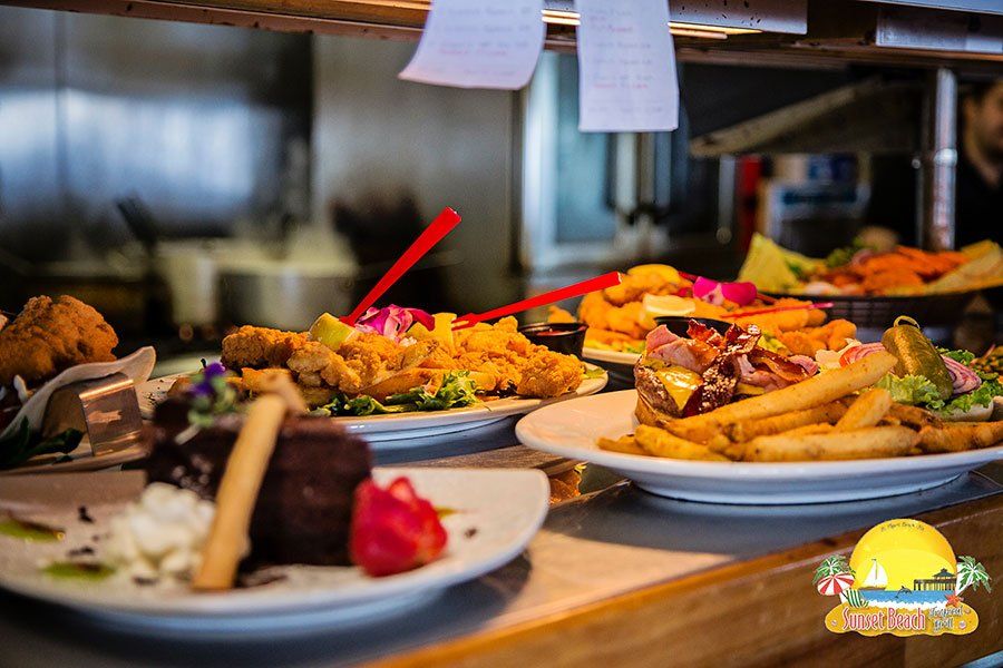 Plated restaurant food: fried chicken, burger, dessert, and vegetables on a countertop near a kitchen.