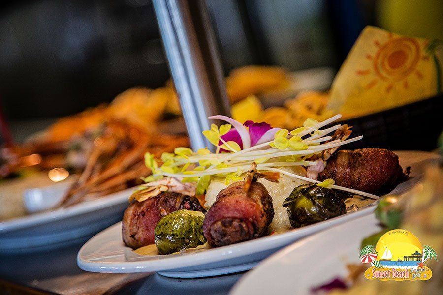 Plate of food: bacon-wrapped bites, sprouts, garnished with flowers, near a lobster and drawing of a sun.