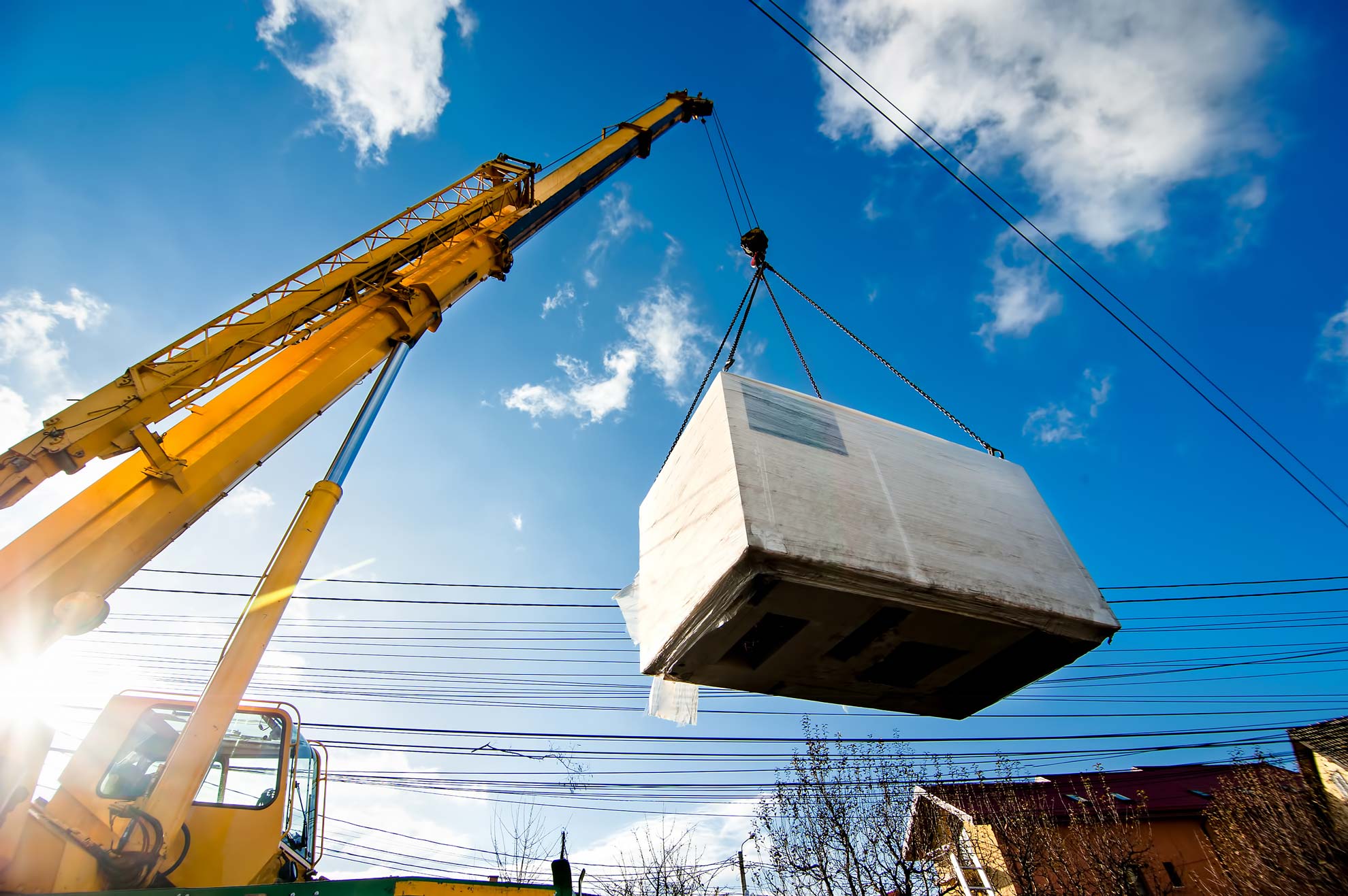 Une grue soulève un grand objet rectangulaire emballé, se détachant sur un ciel bleu avec quelques bâtiments.