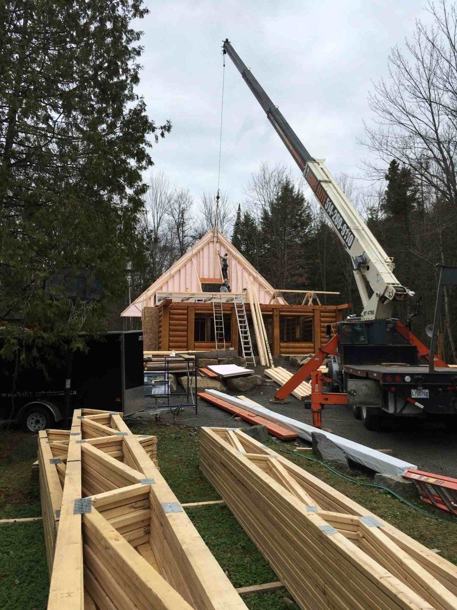 Chantier de construction avec une grue soulevant une poutre de toit sur une cabane en rondins ; fermes en bois au premier plan.