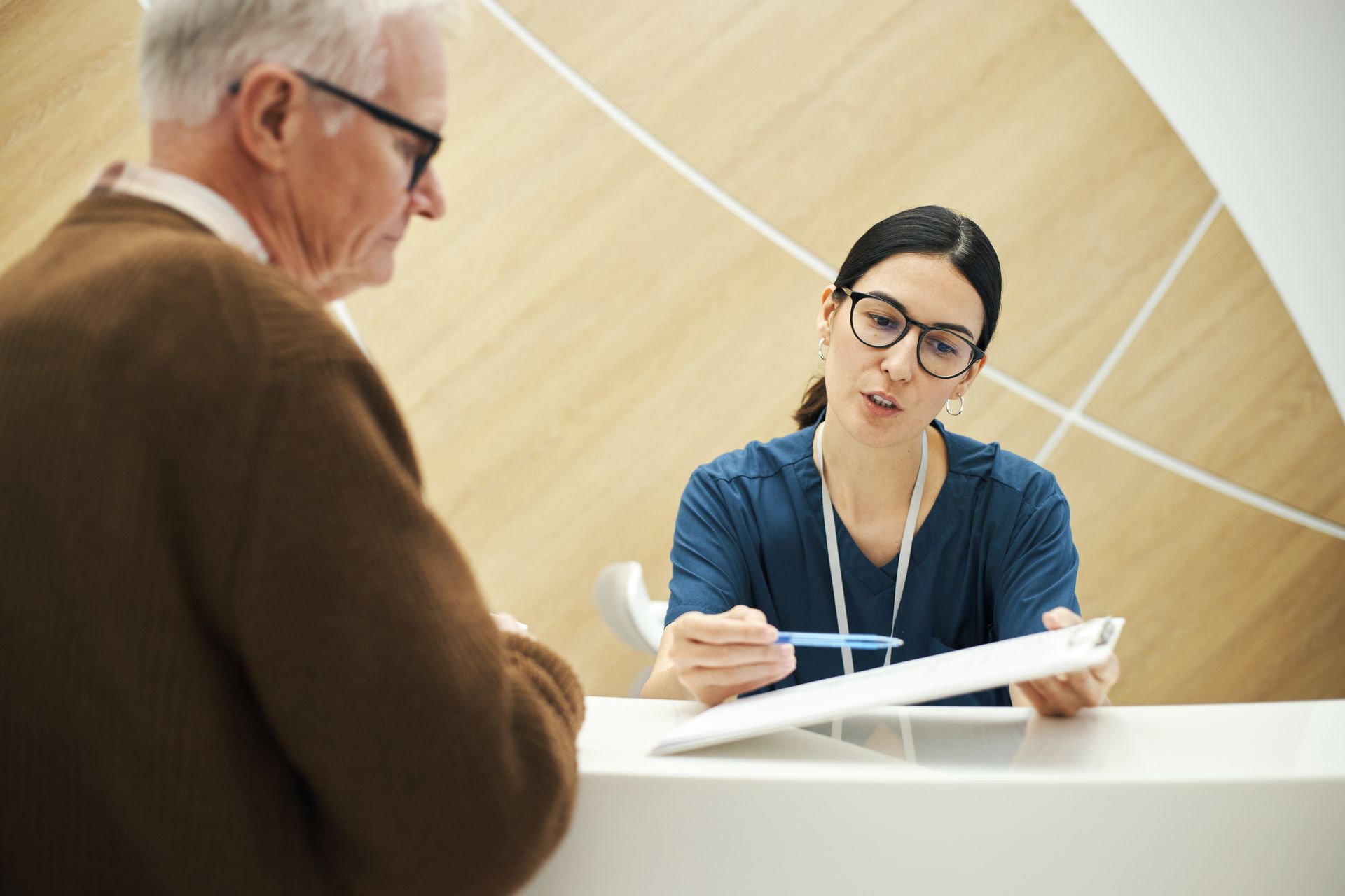 A person in medical scrubs assists a patient at a reception desk, holding a clipboard and pen.