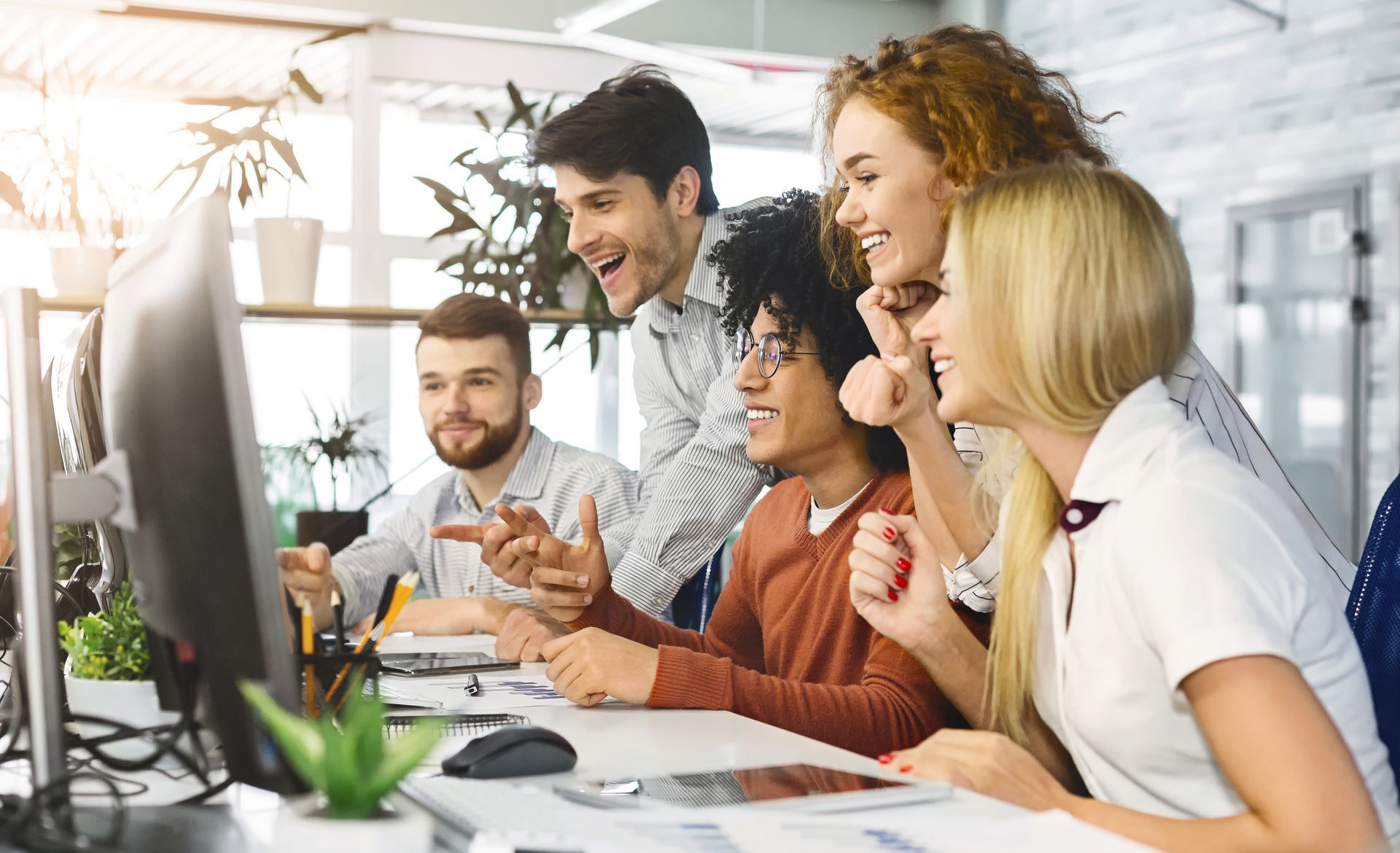 Four colleagues looking at a computer screen in a bright office, smiling and pointing while collaborating on a project.