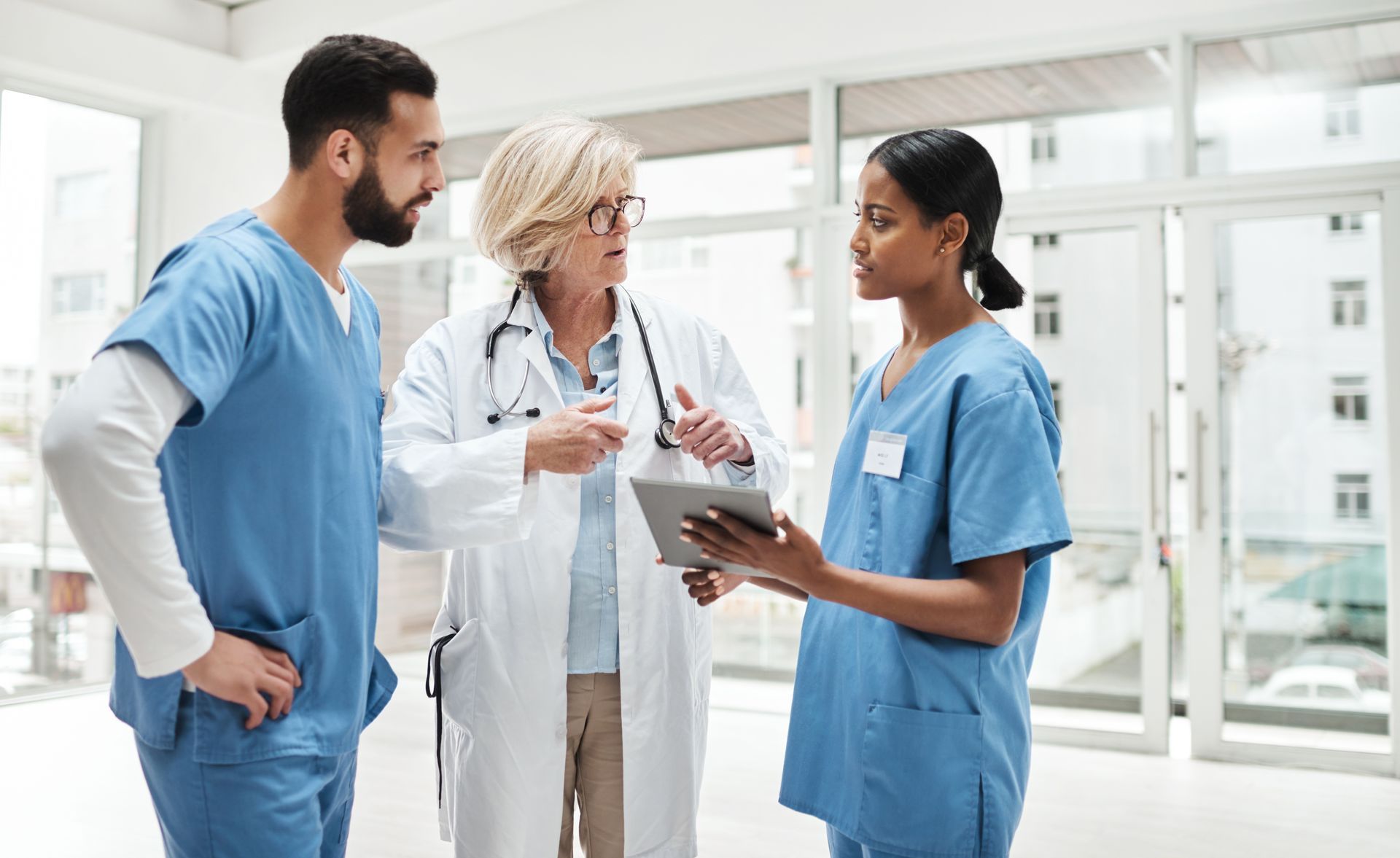 Two medical staff in blue scrubs and a physician in a white coat consult while looking at a tablet in a bright hospital.