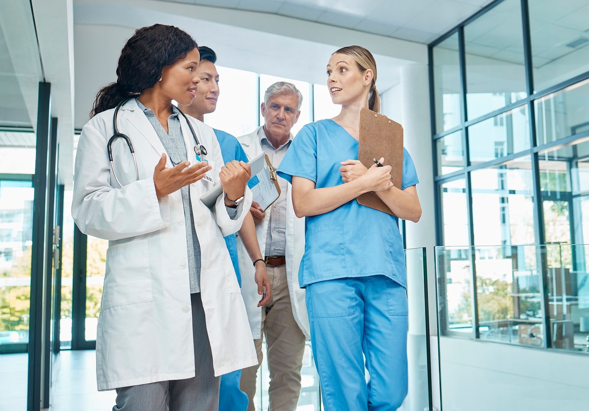 Four medical professionals in scrubs and lab coats walk through a bright, modern hospital hallway, talking to each other.