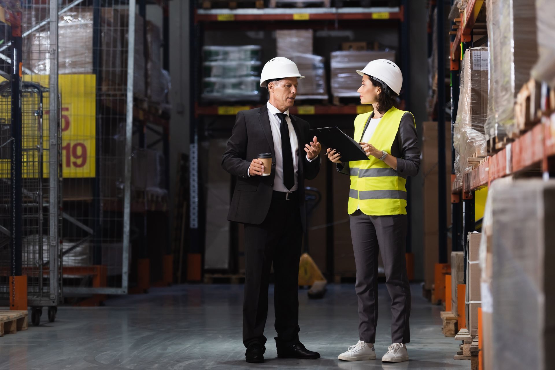 Two professionals in hard hats discuss inventory while standing in a warehouse aisle with shelves of goods.
