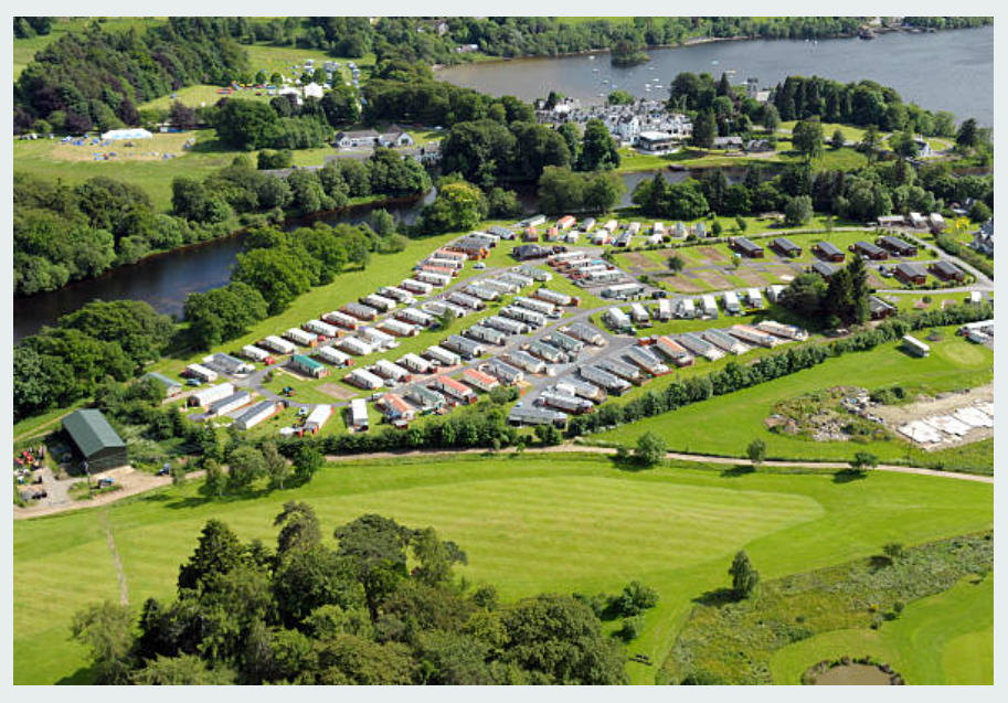 Aerial view of a caravan park with rows of caravans, green fields, trees, and a lake in the background.
